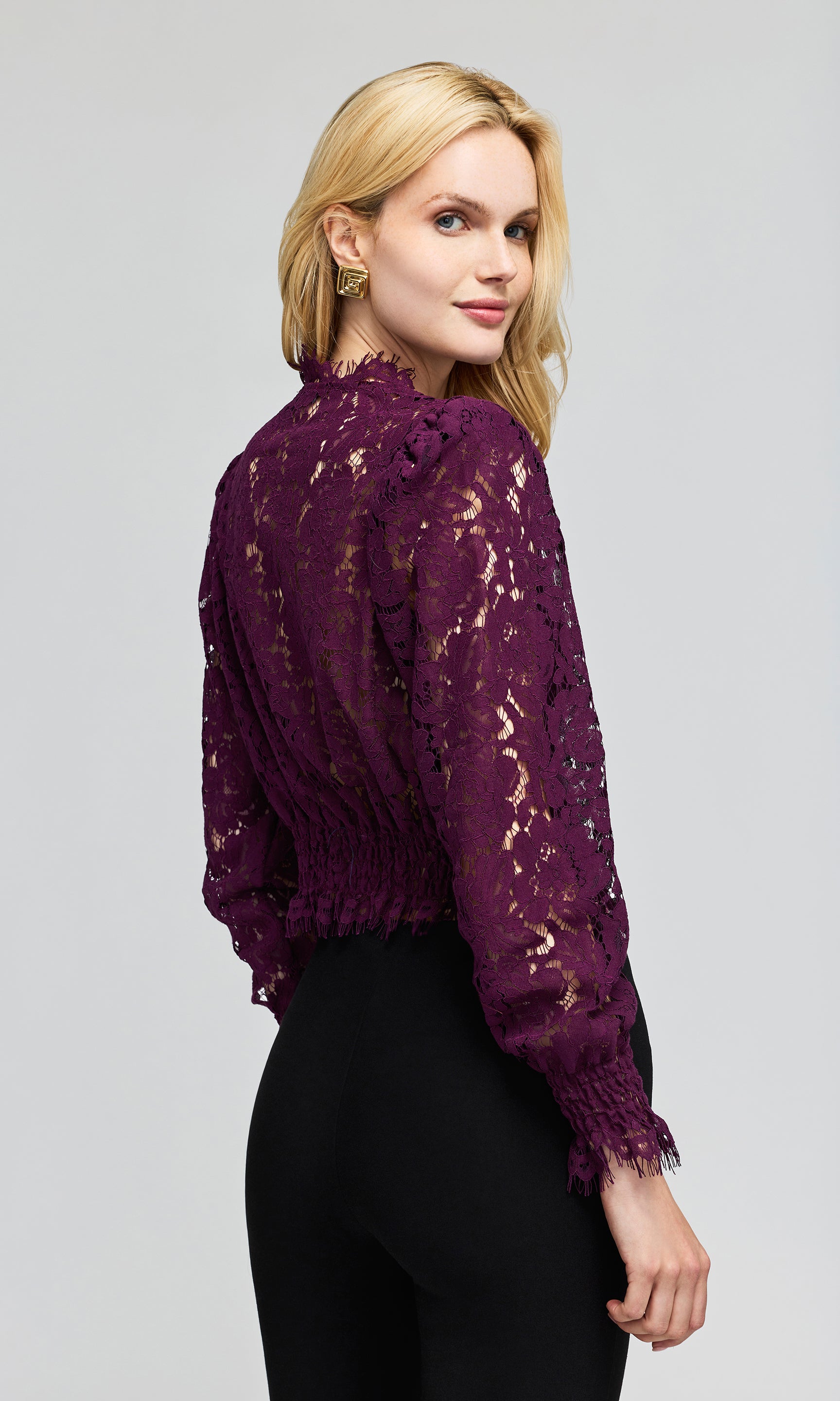 Side view of model wearing a deep purple lace long-sleeve blouse with a deep V-neck and black pants, on a plain background.
