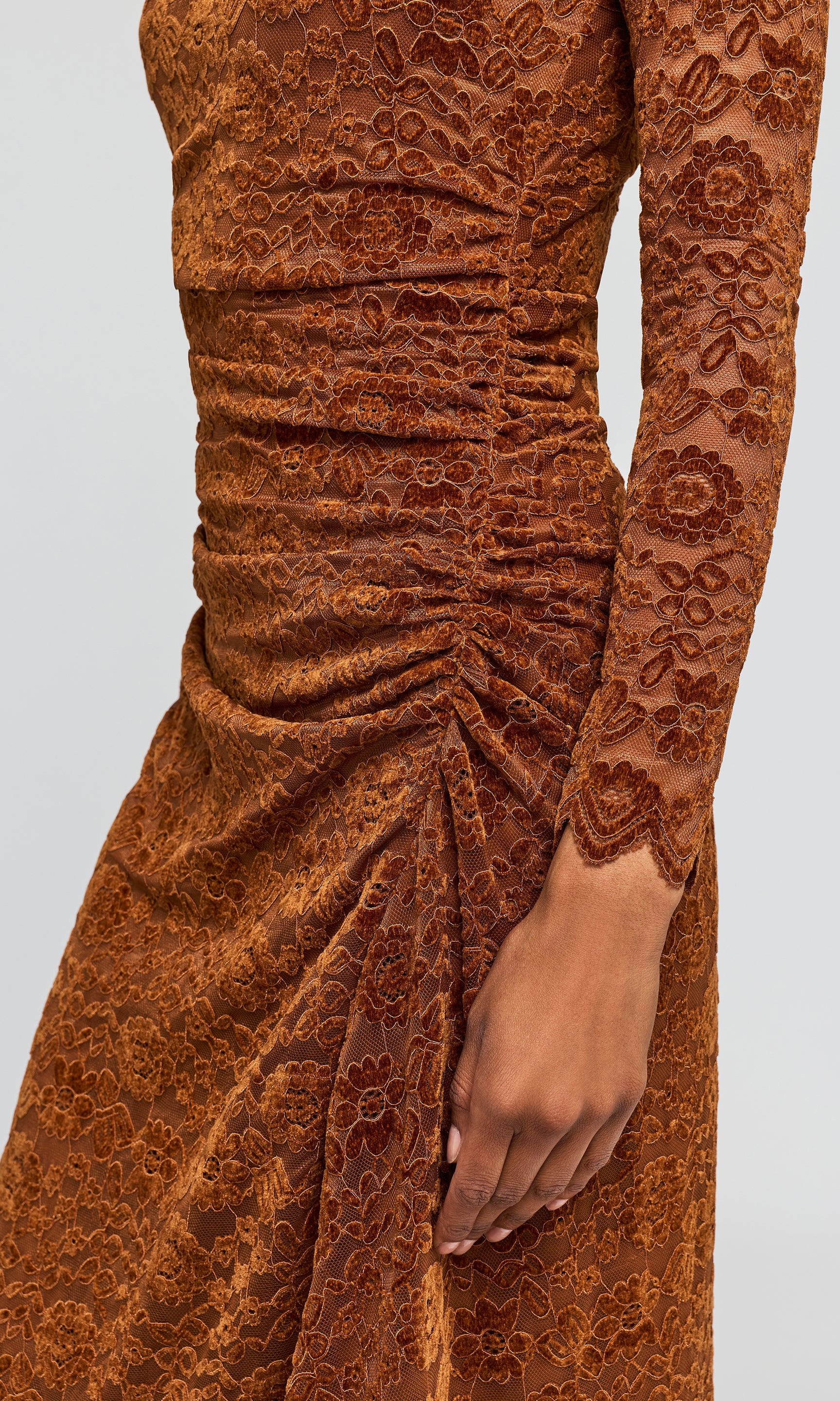 A close-up of a long-sleeve ruched dress in a warm rust-brown color, featuring a textured floral lace overlay and gathered detailing at the waist.