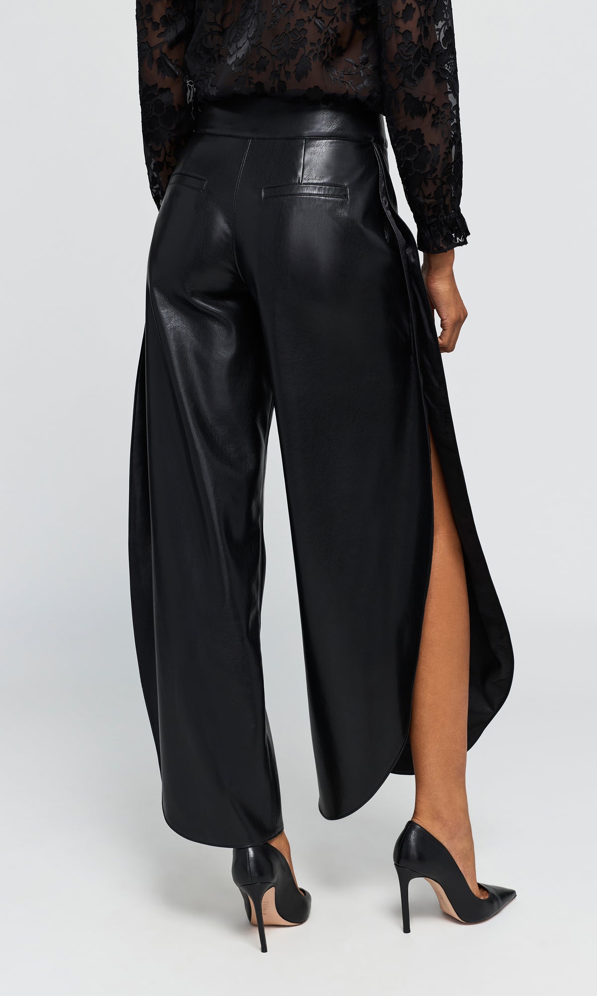Back view of model wearing black wide-leg trousers with a glossy faux-leather finish, paired with a black lace long-sleeve top and black pointed-toe heels.