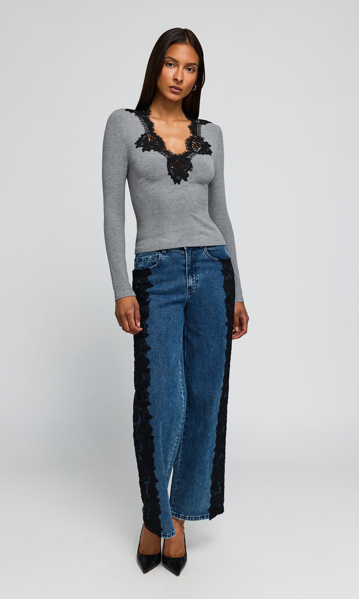 Model wearing a gray long-sleeve knit top with black lace trim at the neckline, paired with wide-leg blue jeans featuring black lace side panels, and black pointed heels.