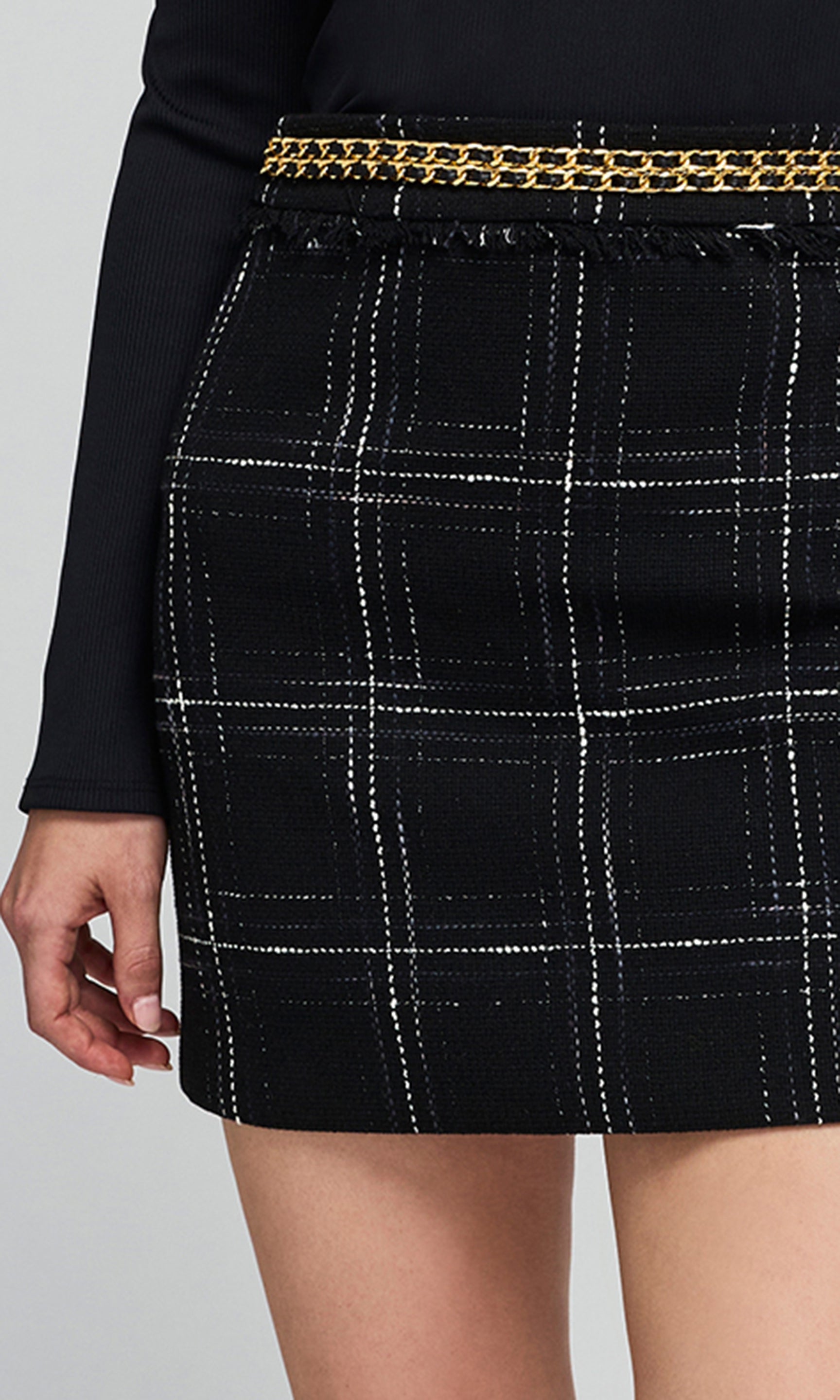 Close-up of a model wearing a black tweed mini skirt with a white windowpane plaid pattern and a gold chain belt at the waist.