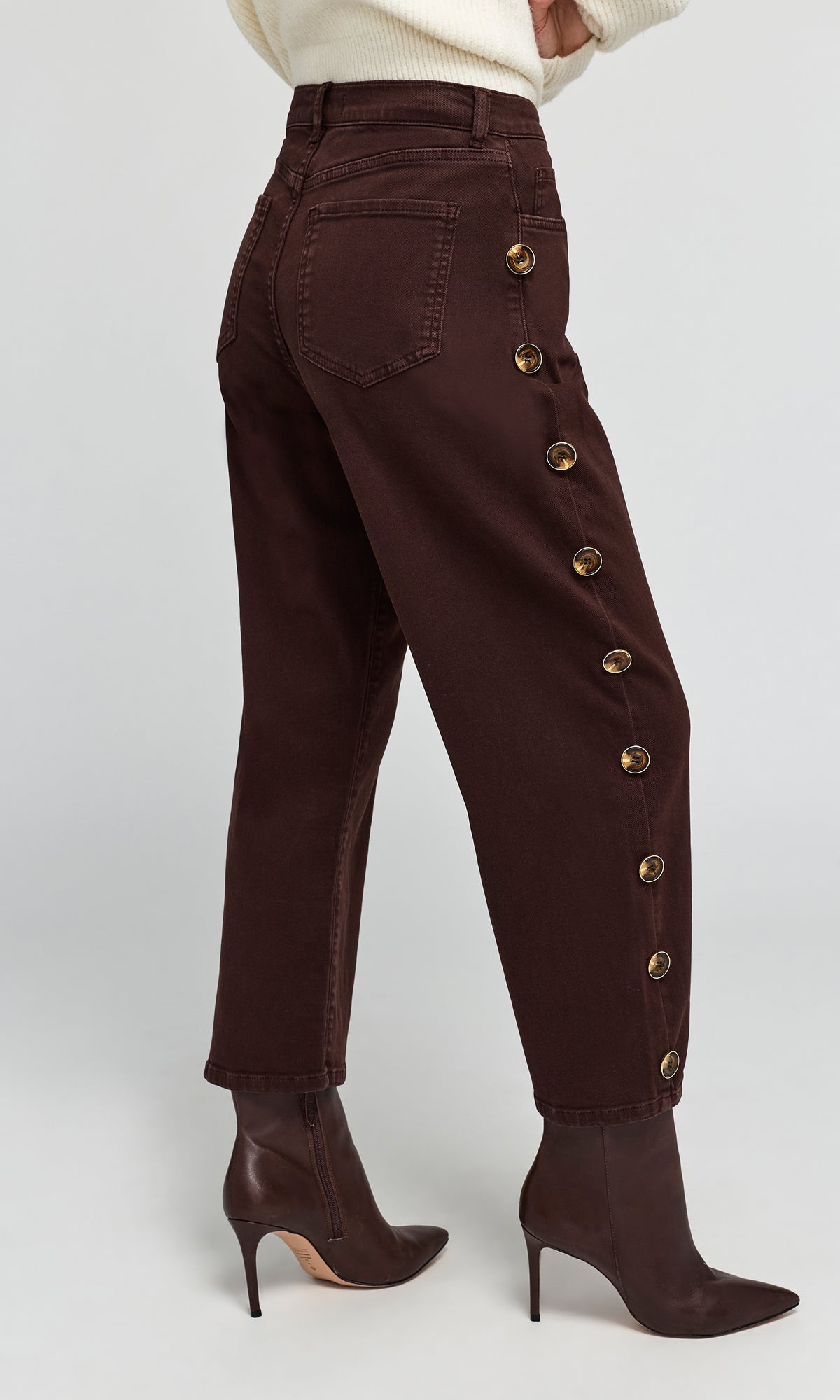 3/4 back view of model wearing dark brown cropped jeans with decorative gold-tone side buttons, paired with a cream sweater.