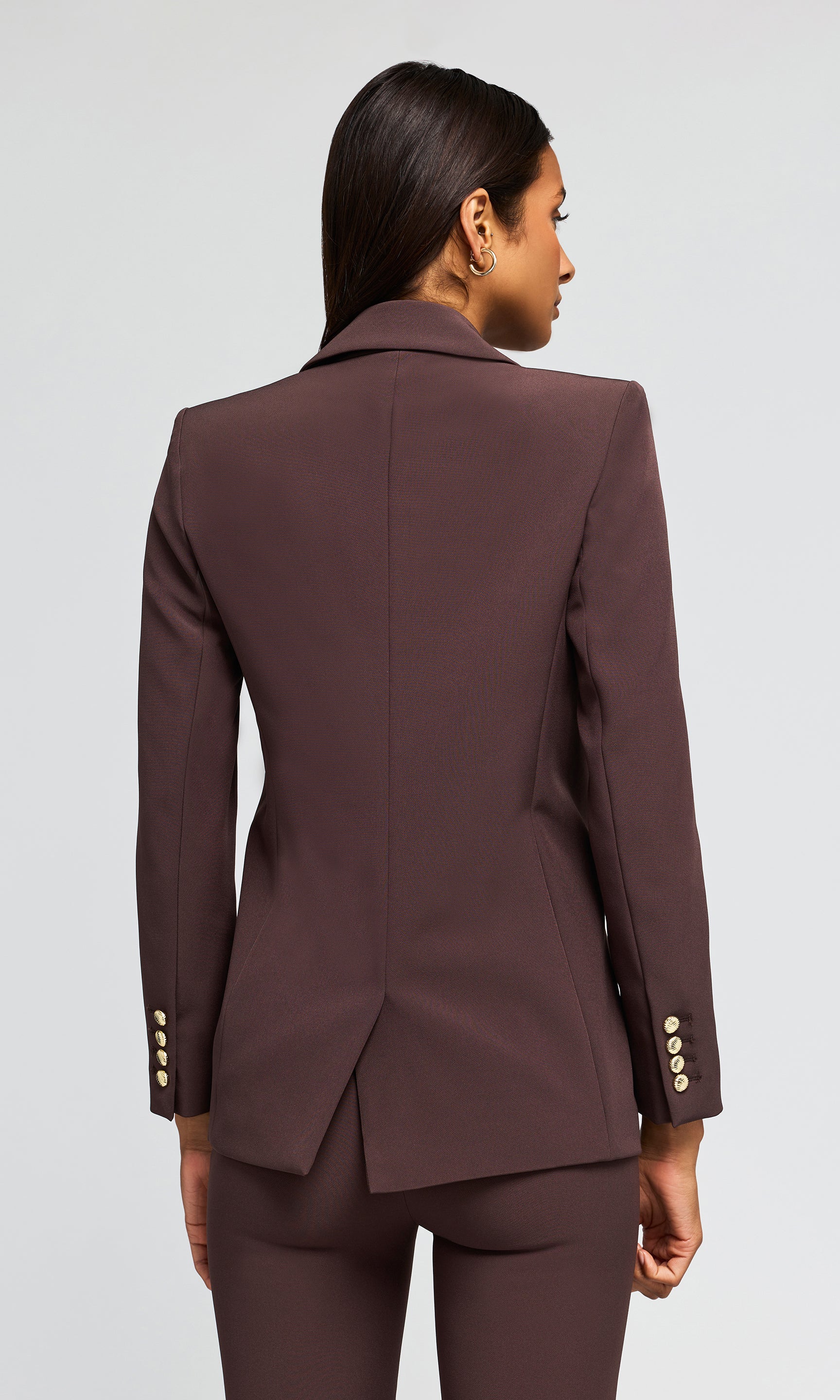 Back view of model wearing a dark brown double-breasted blazer with gold buttons and matching trousers.