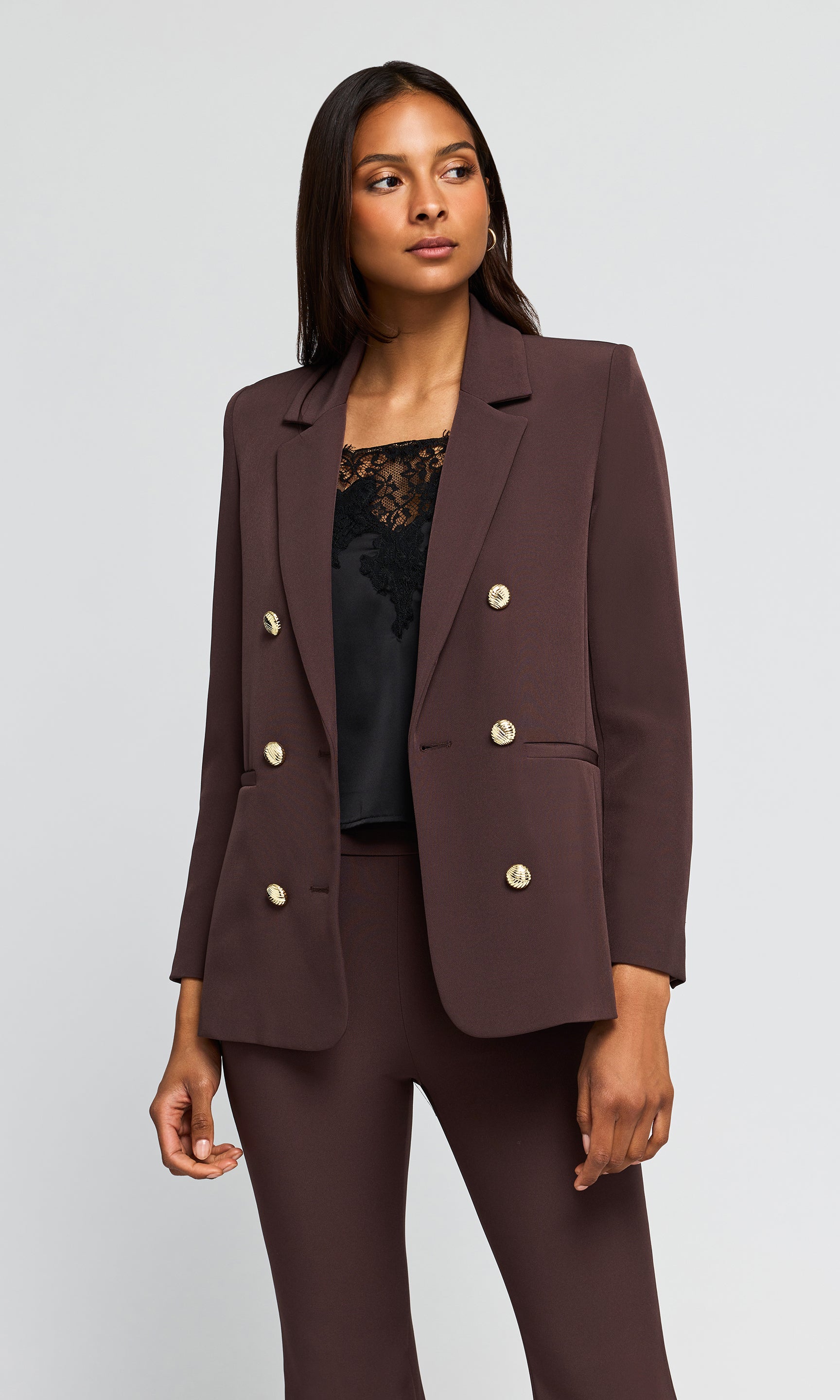 Side view of model wearing a dark brown double-breasted blazer with gold buttons and matching trousers.