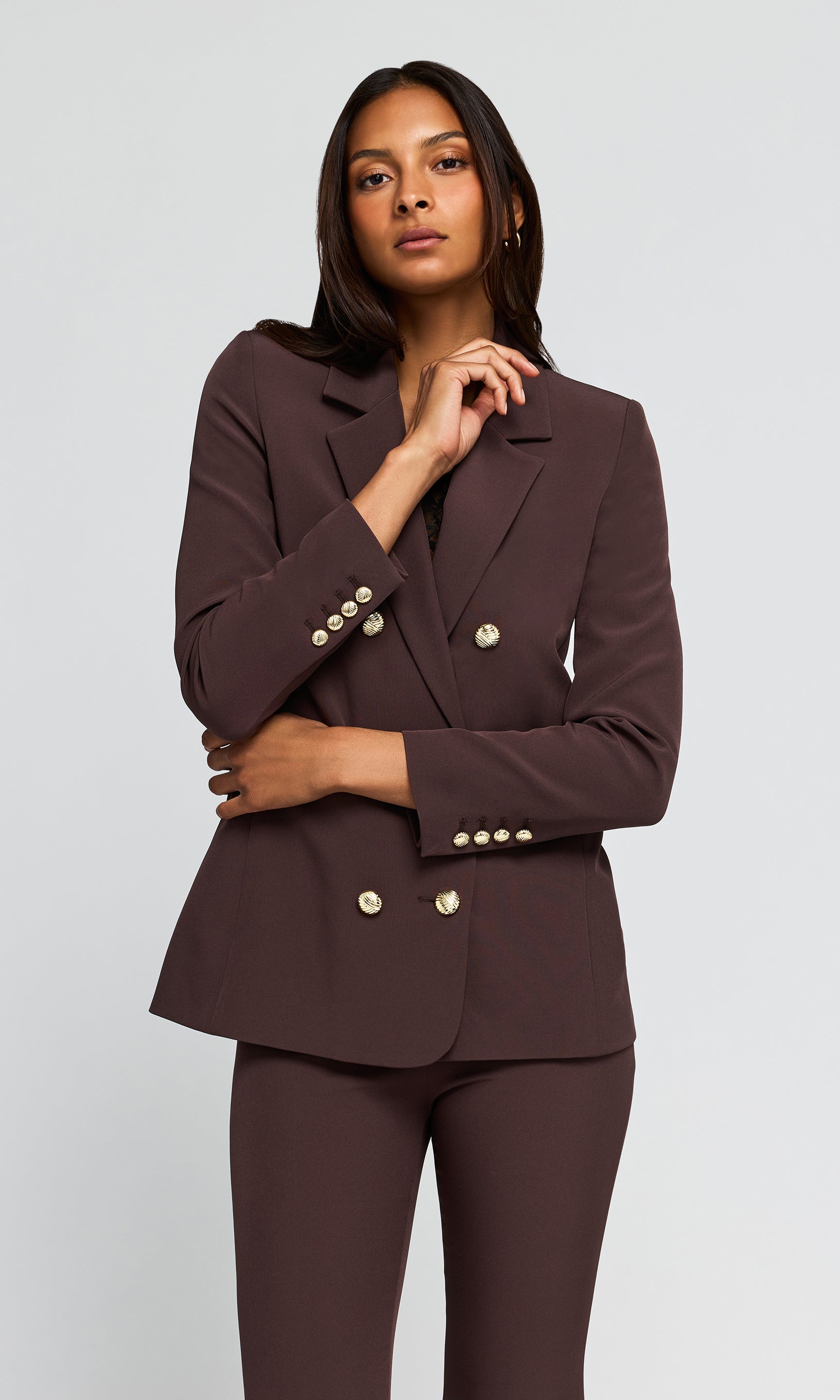 A woman modeling a dark brown double-breasted blazer with gold buttons and matching dark brown trousers against a light gray studio background.
