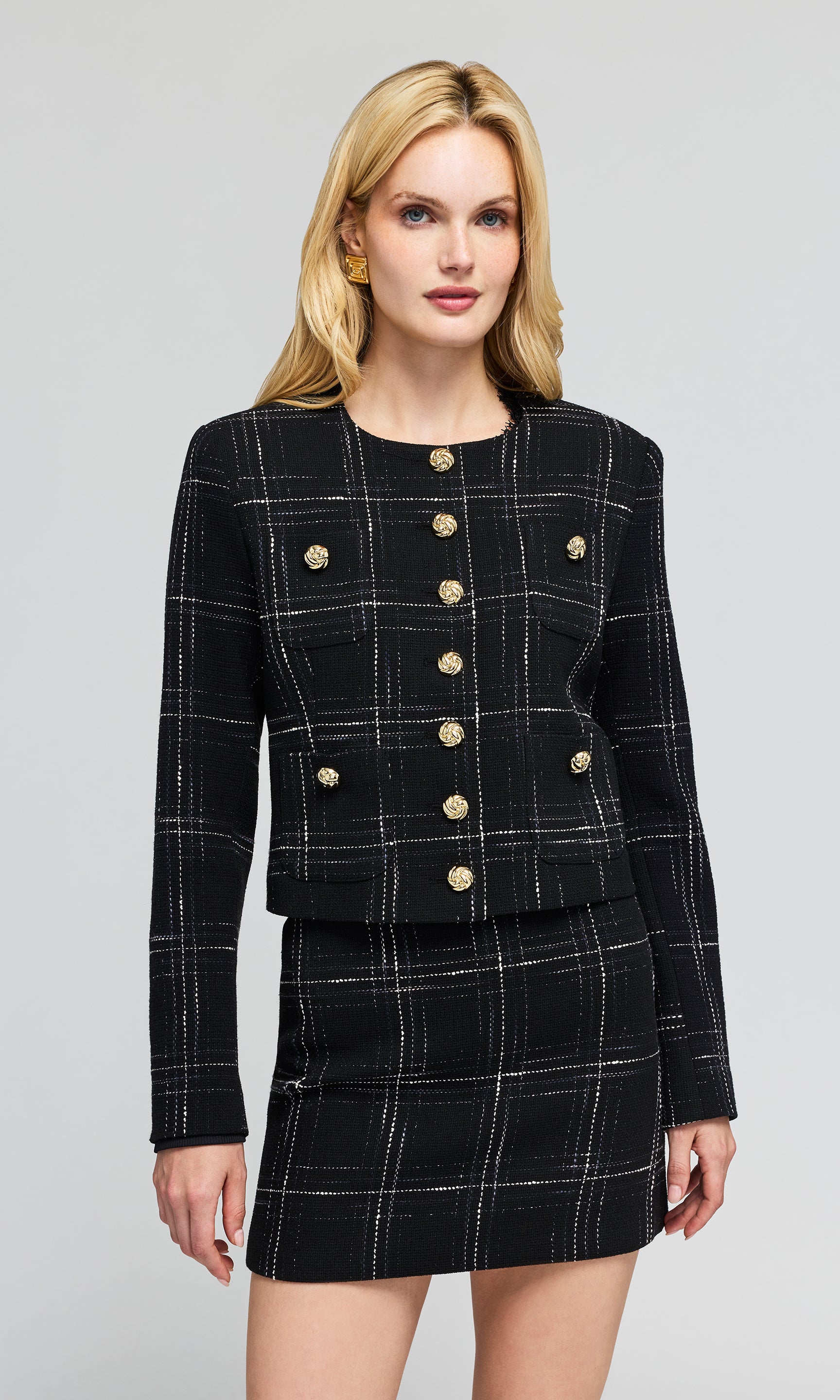 Model wearing a black tweed blazer and matching mini skirt set with a white windowpane plaid pattern and prominent gold button.