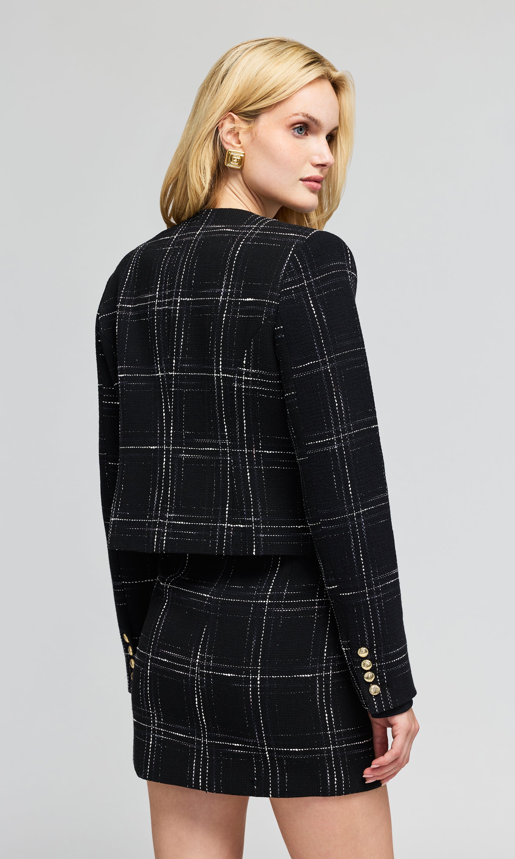 Back view of model wearing a black plaid blazer and matching mini skirt with white windowpane lines.