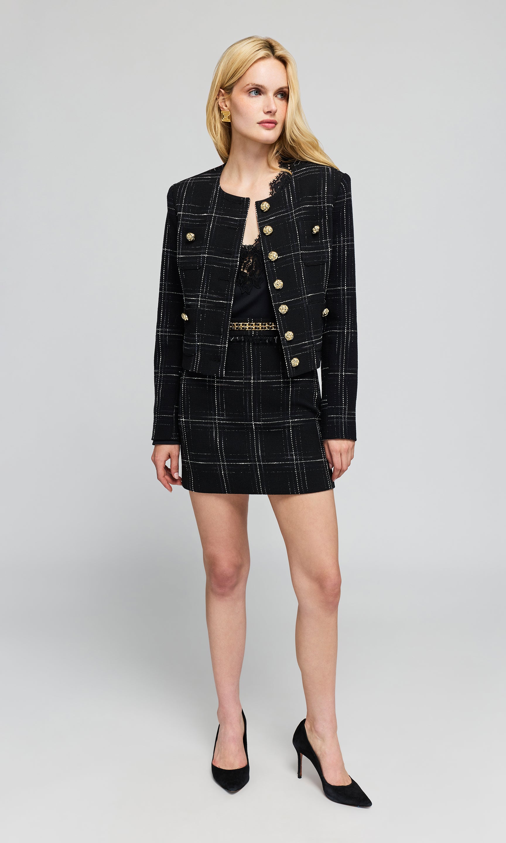 Model wearing a black double-breasted plaid blazer and matching mini skirt with gold buttons, paired with a black top and black high heels.