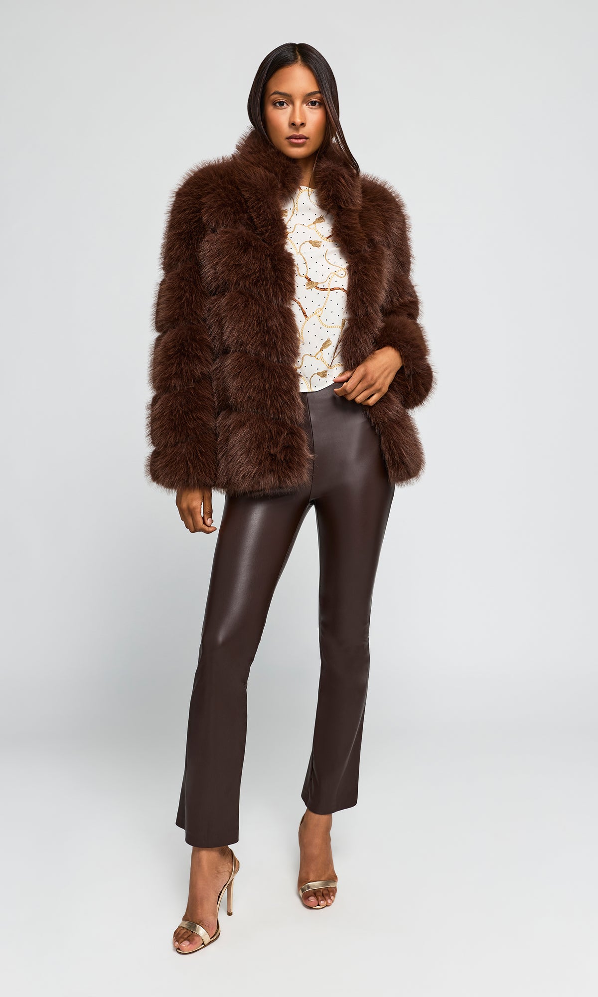 Model wearing a dark brown shaggy faux-fur jacket over a cream patterned top, dark brown faux-leather pants, and metallic heeled sandals.