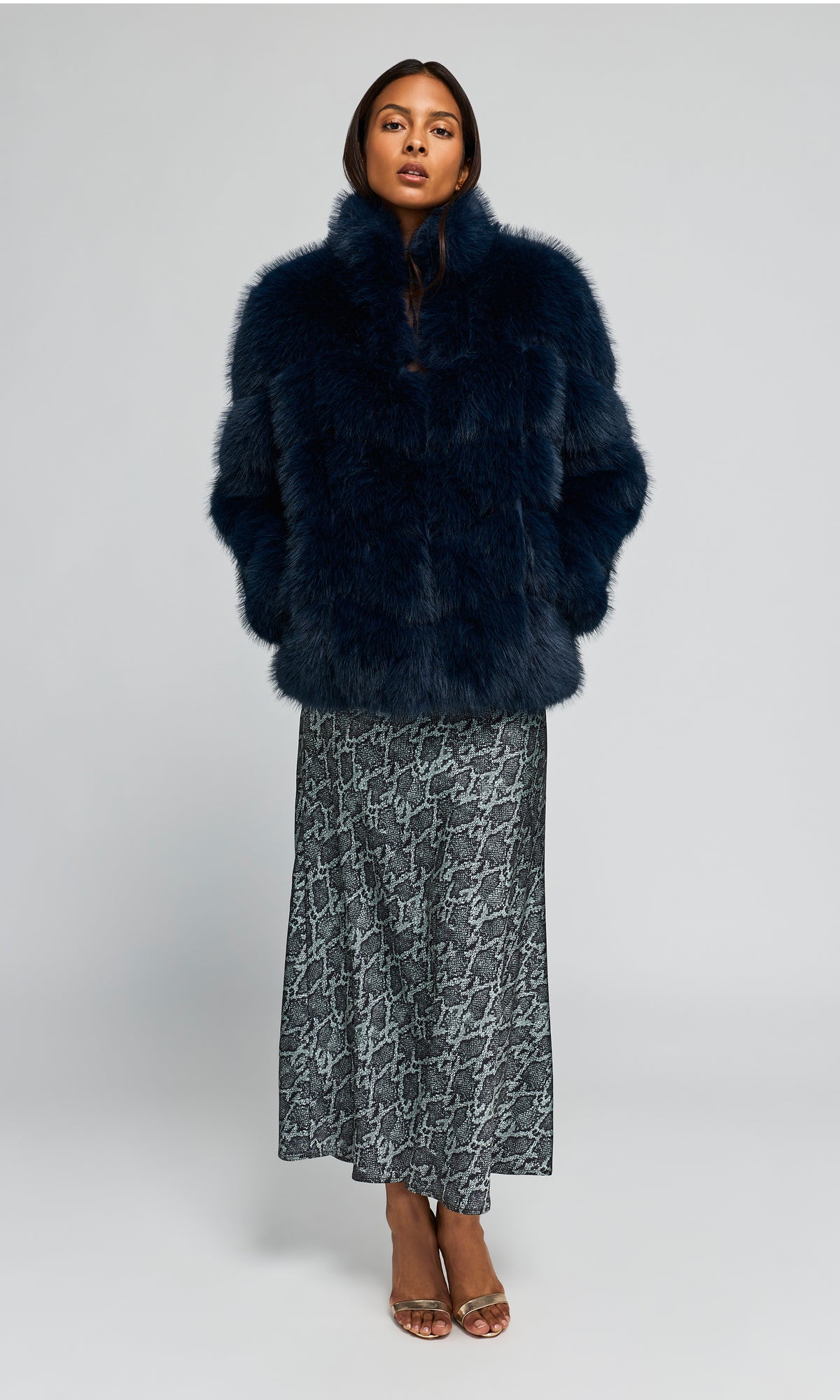 Model wearing a voluminous, dark navy blue faux fur jacket over a snake patterned gray dress against a plain background.