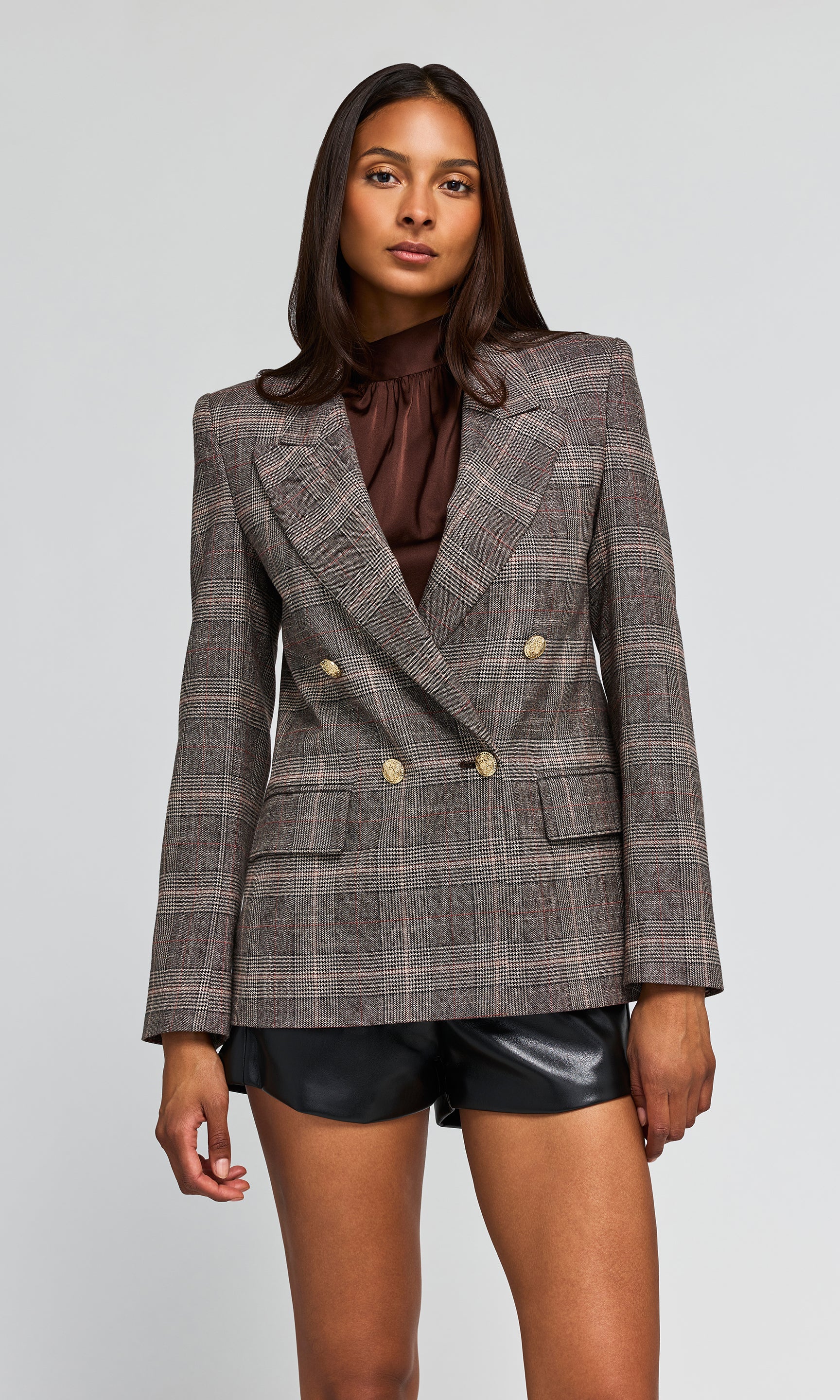 Model wearing a double-breasted plaid blazer in brown and gray tones, paired with a dark brown ruffled top and black shorts.