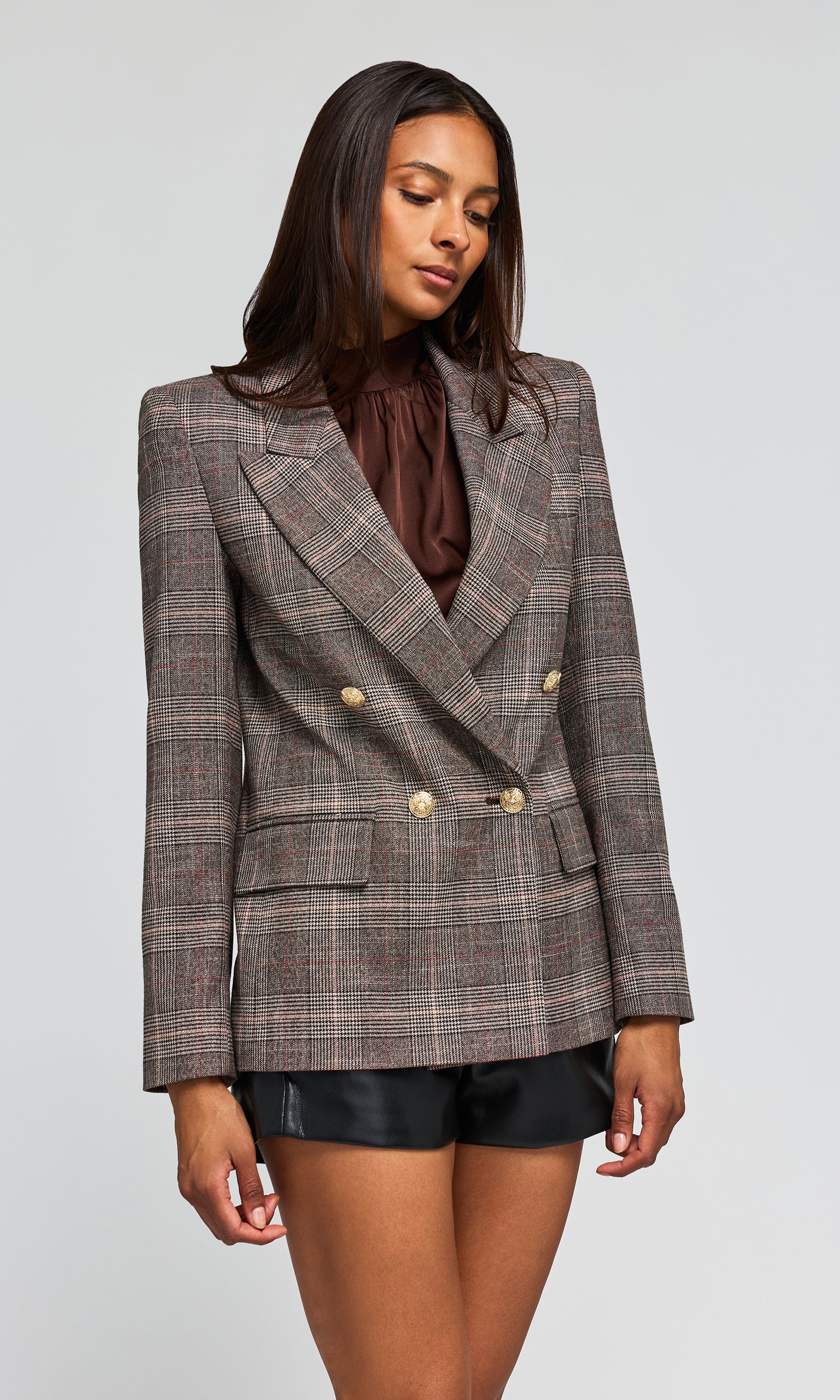 Side view of model wearing a double-breasted plaid blazer in brown and gray tones, paired with a dark brown ruffled top and black shorts.
