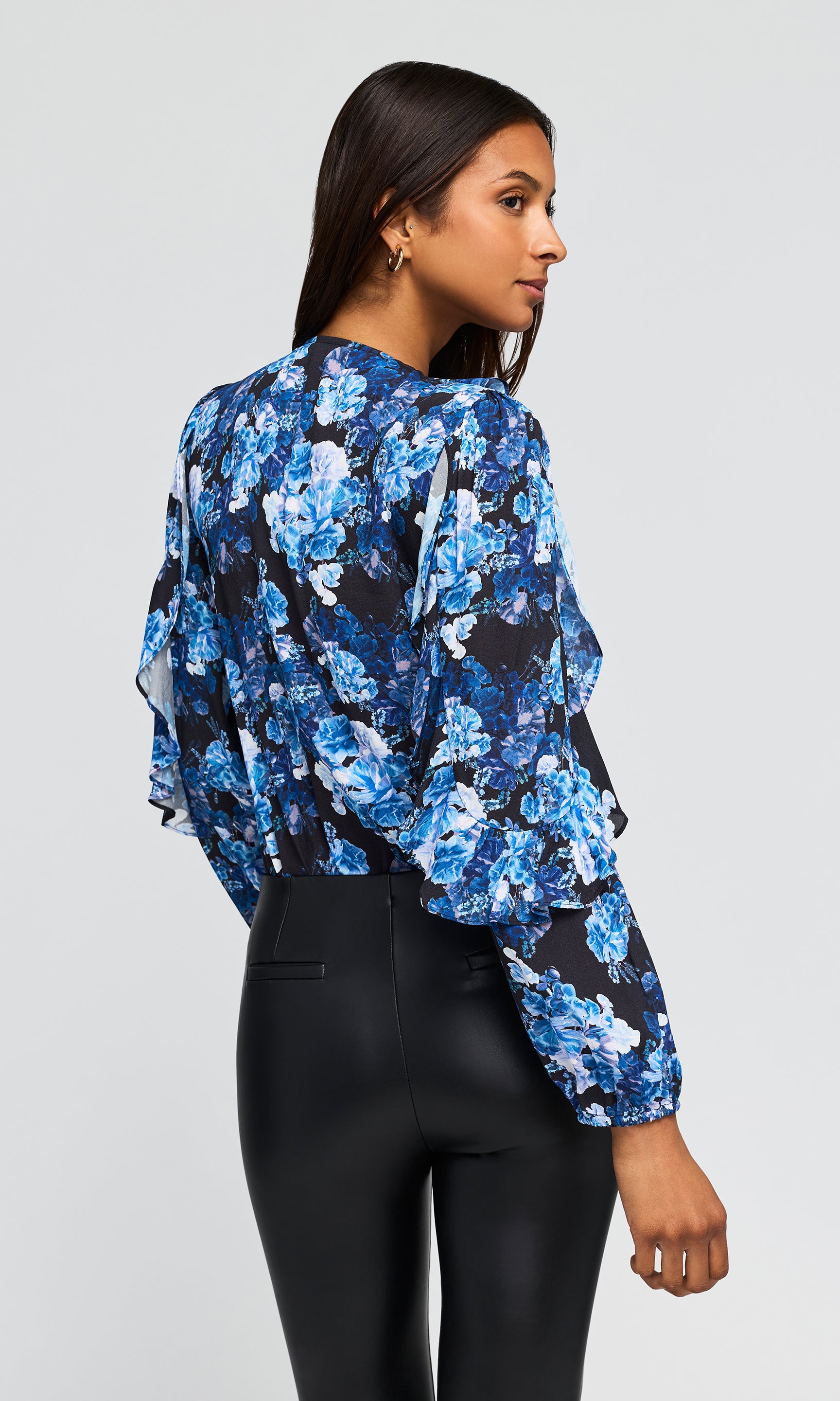 Back view of model wearing a floral blouse with a deep V-neck, featuring blue and white flowers, paired with black faux-leather pants.