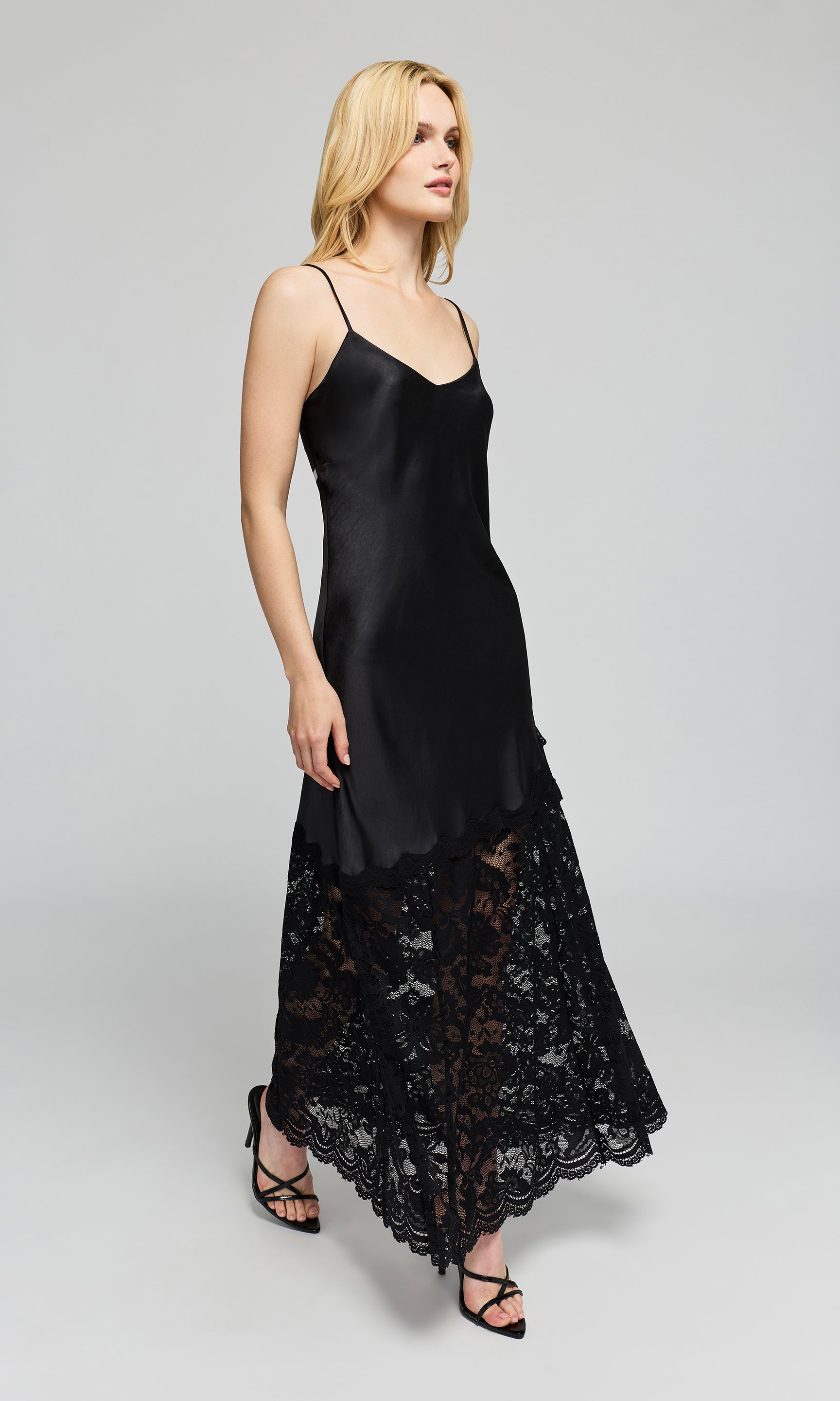 Side view of model wearing a black satin slip dress with thin straps and a black lace hem, on a plain background.
