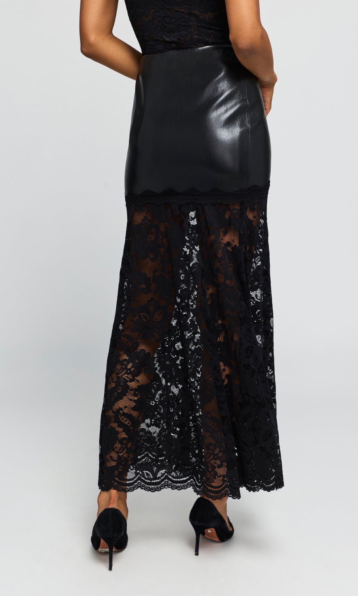 Back view of model wearing a black midi skirt featuring a smooth faux-leather upper section and a sheer black lace lower skirt.