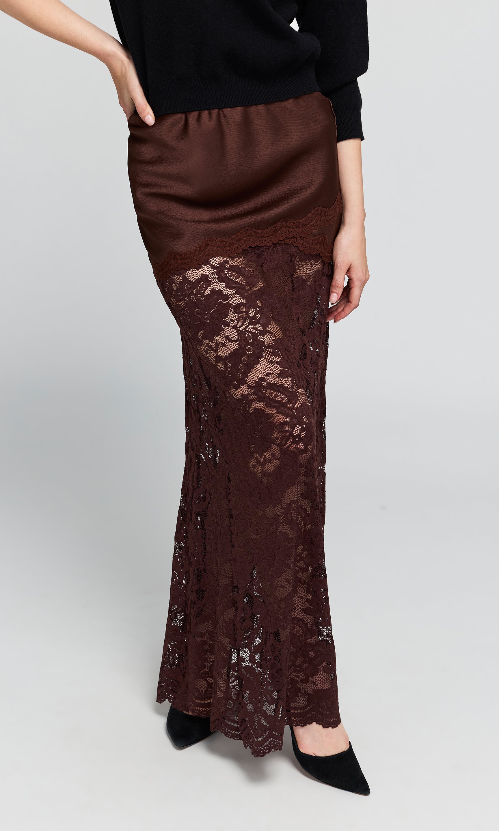 A woman wearing a long brown satin and lace skirt with black pointed-toe heels, shown from the waist down.