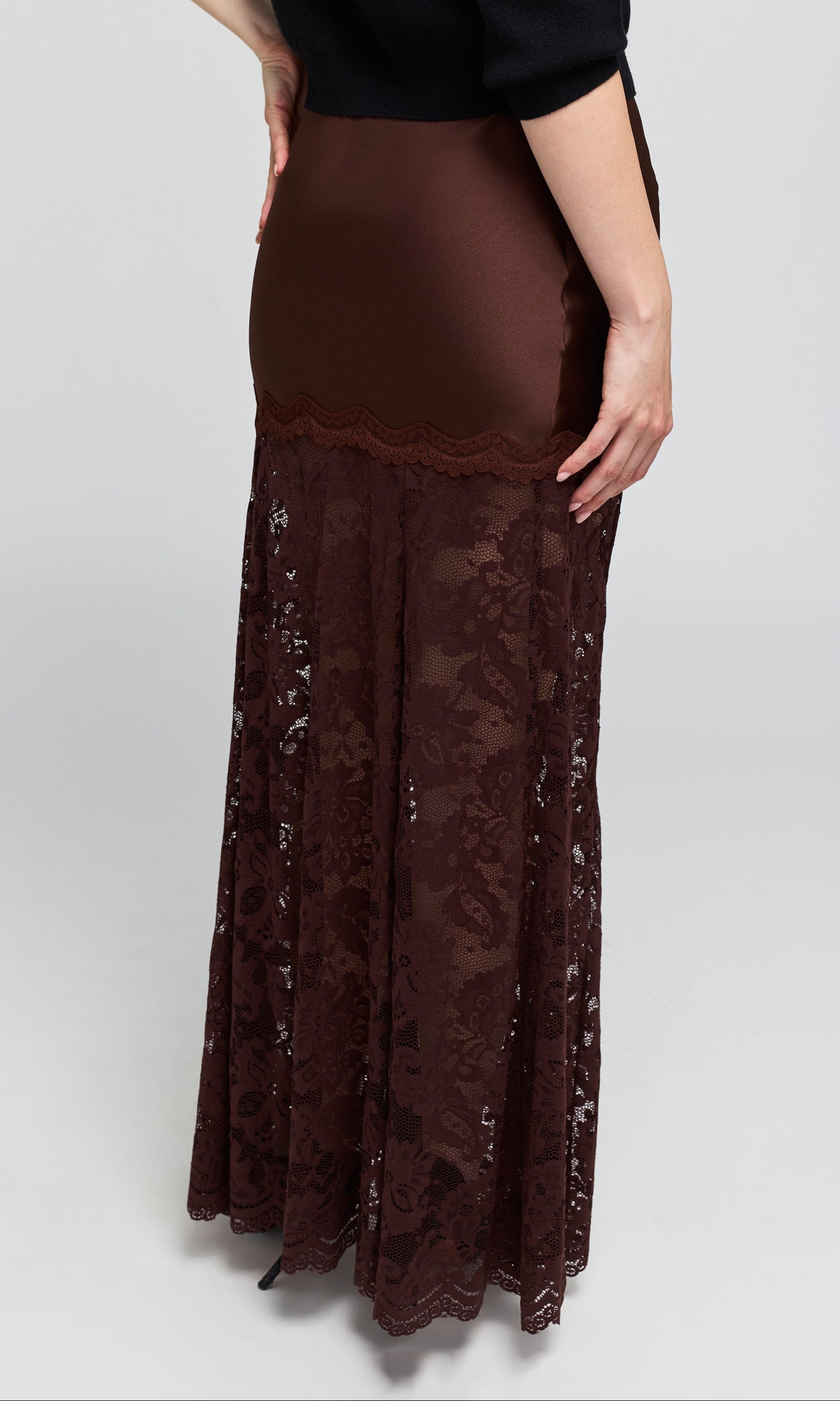 Back view of a dark brown lace maxi skirt worn by a model, styled with a black long-sleeve top and black pointed-toe heels.