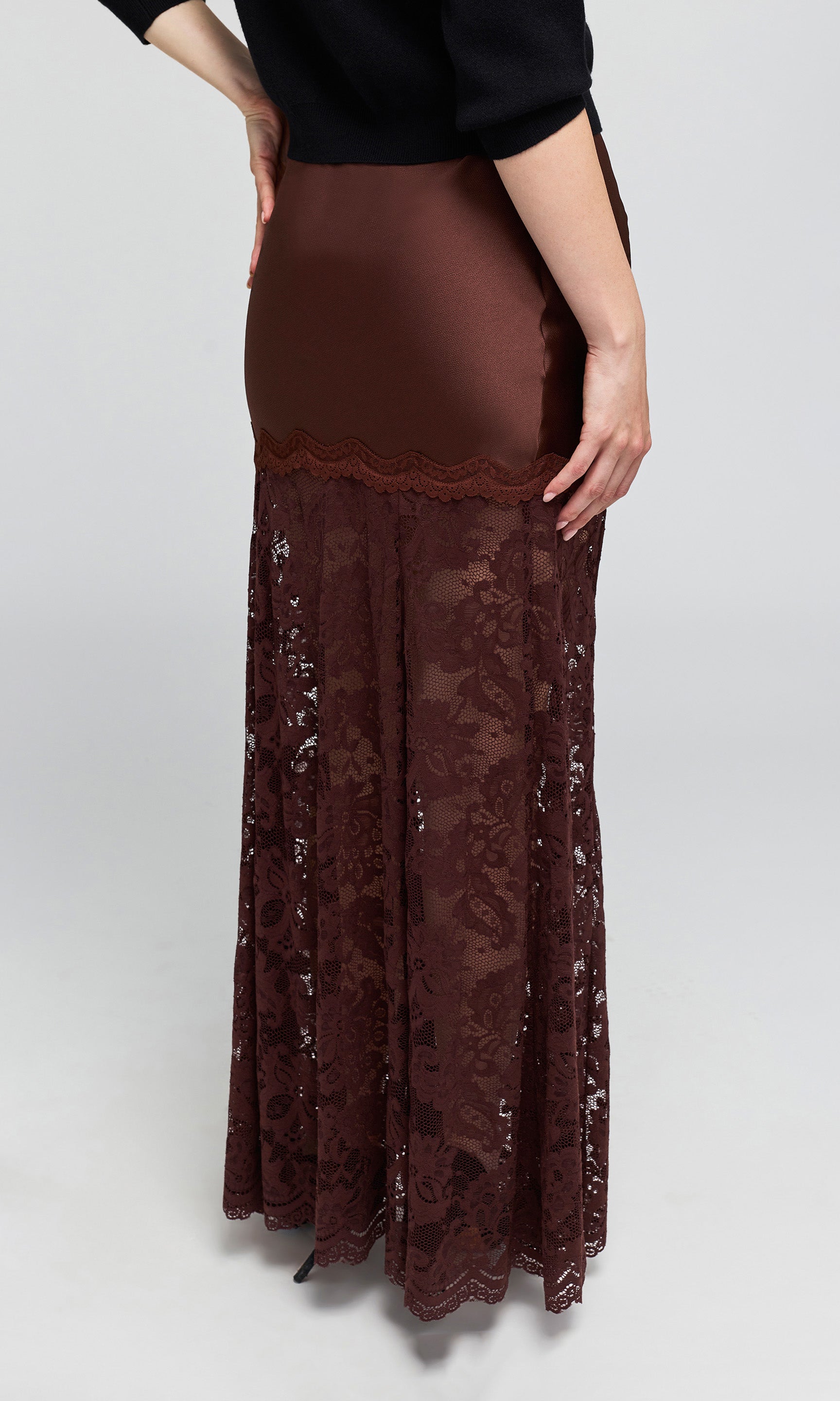 Back view of model wearing a brown lace maxi skirt with a sheer satin-like fabric, worn by a model with a black top.