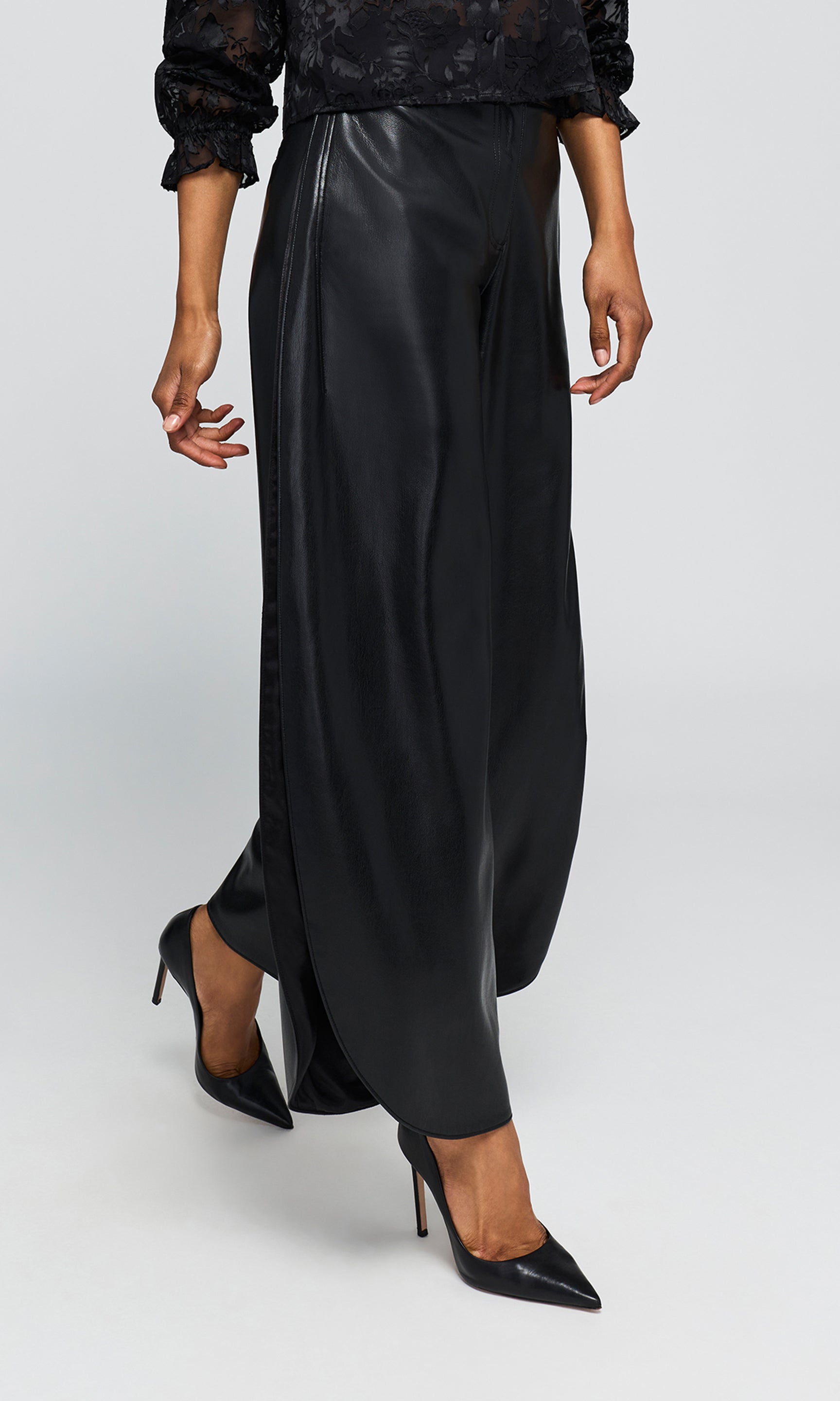 Side view of model wearing black wide-leg trousers with a glossy faux-leather finish, paired with a black lace long-sleeve top and black pointed-toe heels.