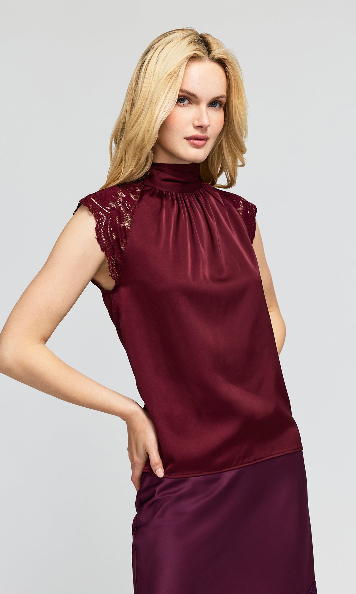 Side view of model wearing a sleeveless high-neck turtleneck blouse in deep burgundy with lace cap sleeves and a matching skirt.