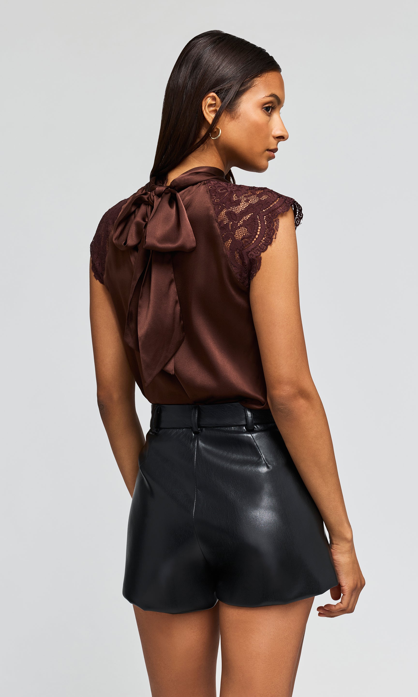 A woman shown from the back wearing a dark brown satin top with a large bow and lace shoulder detail, paired with black faux-leather shorts.