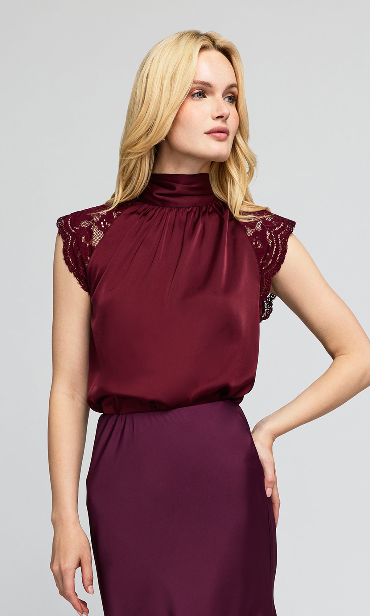 Model wearing a sleeveless high-neck turtleneck blouse in deep burgundy with lace cap sleeves and a matching skirt.