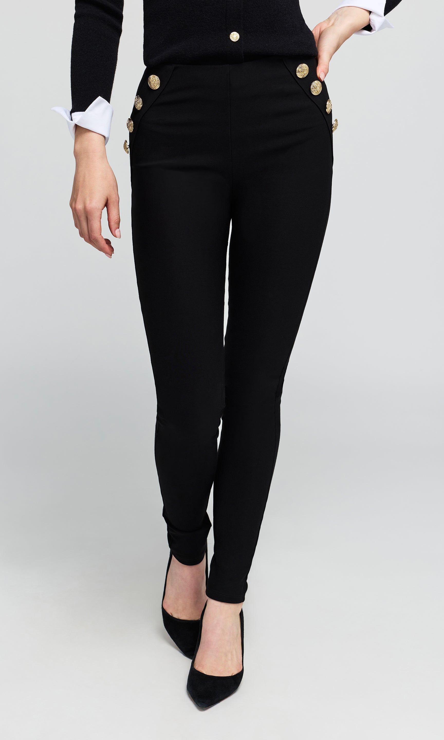 Black skinny pants with decorative gold buttons on the hips, shown on a model wearing a black top and black heels against a light gray background.