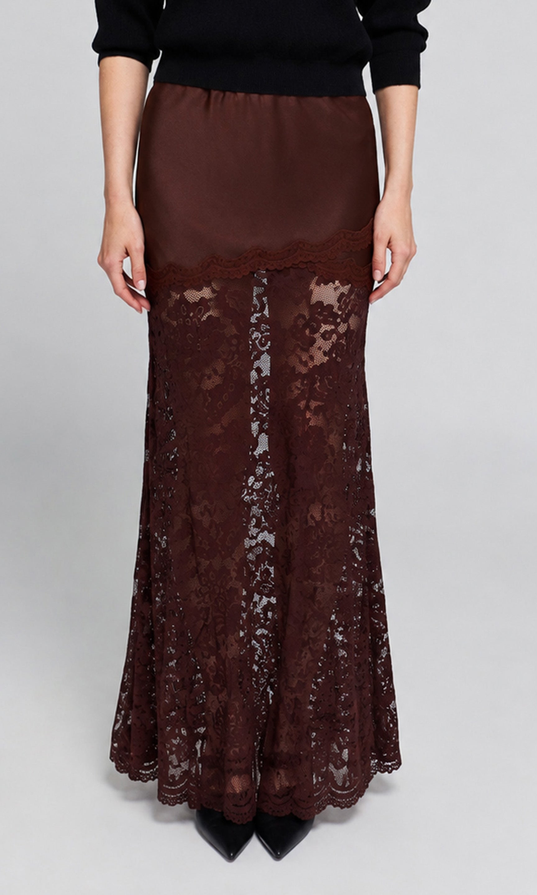 A dark brown lace maxi skirt worn by a model, styled with a black long-sleeve top and black pointed-toe heels.