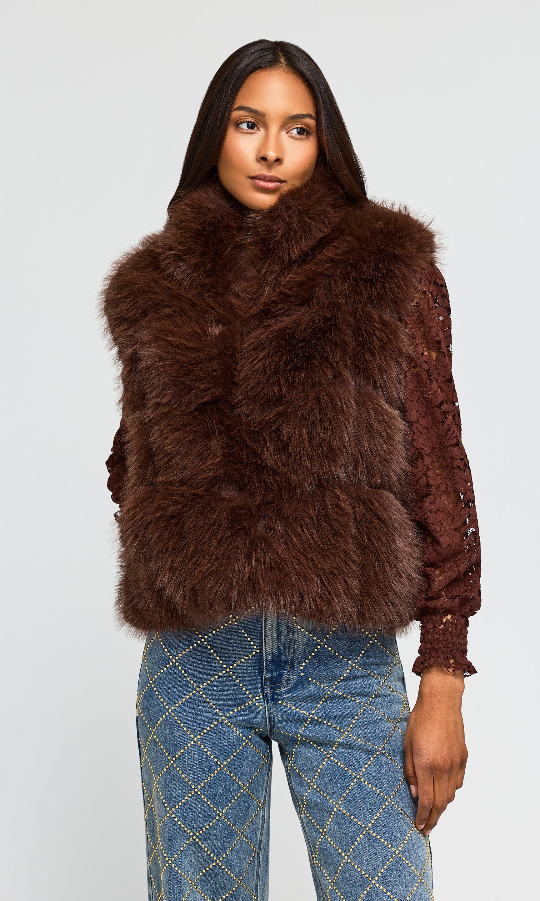 Model wearing a brown faux fur vest with a high collar over a brown lace long-sleeve top and blue jeans.