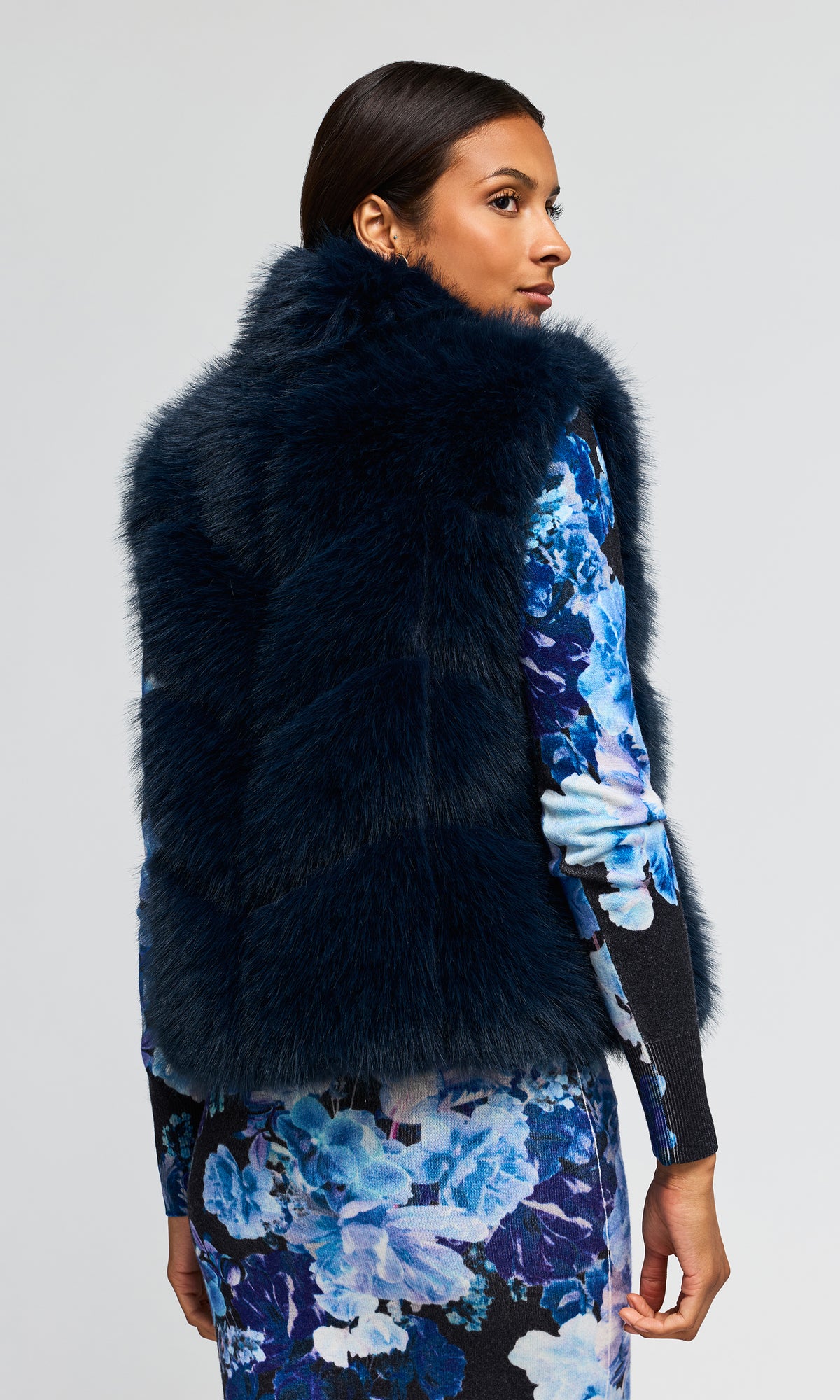 Back view of model wearing a dark navy blue faux fur vest over a blue and black floral long-sleeve cardigan and skirt set.