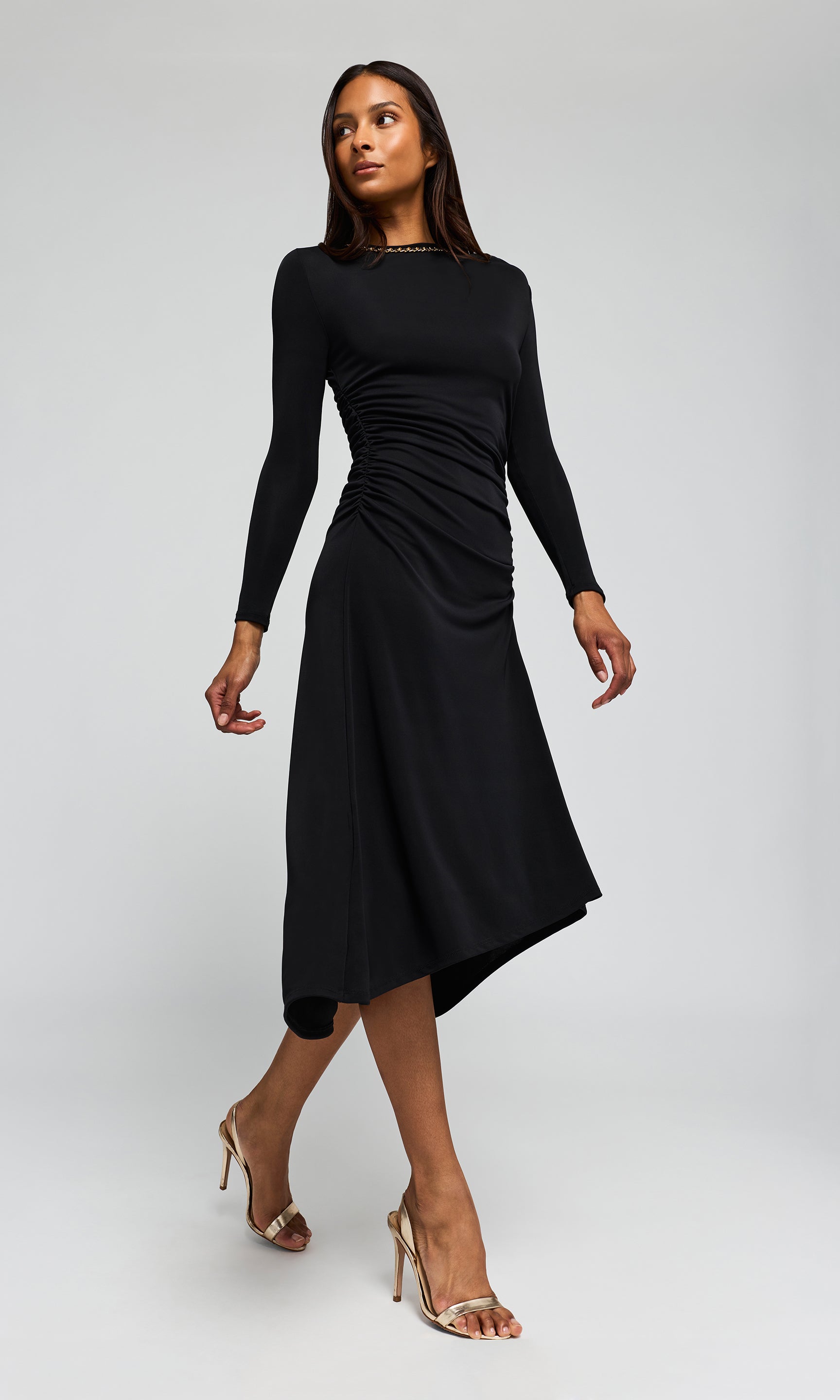 Side view of model wearing a black long-sleeve midi dress with a gathered ruched bodice and asymmetrical draped skirt, on a plain background.