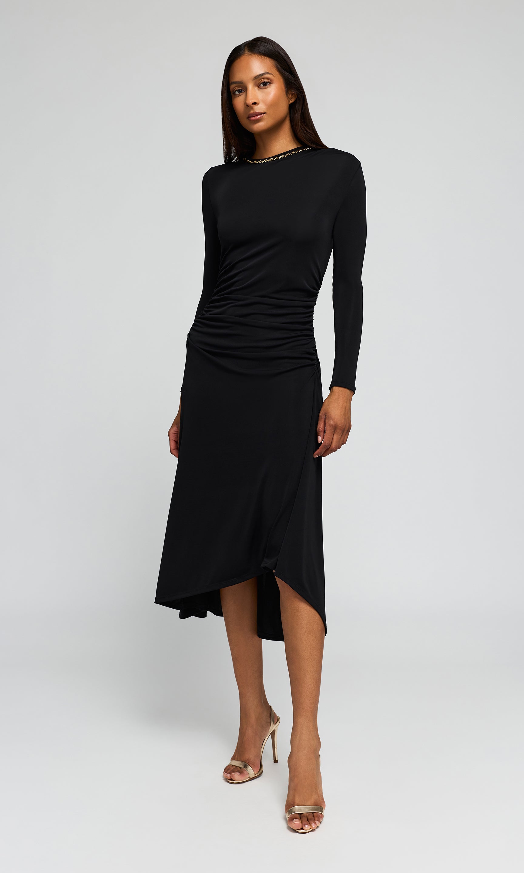 Model wearing a black long-sleeve midi dress with a gathered ruched bodice and asymmetrical draped skirt, on a plain background.