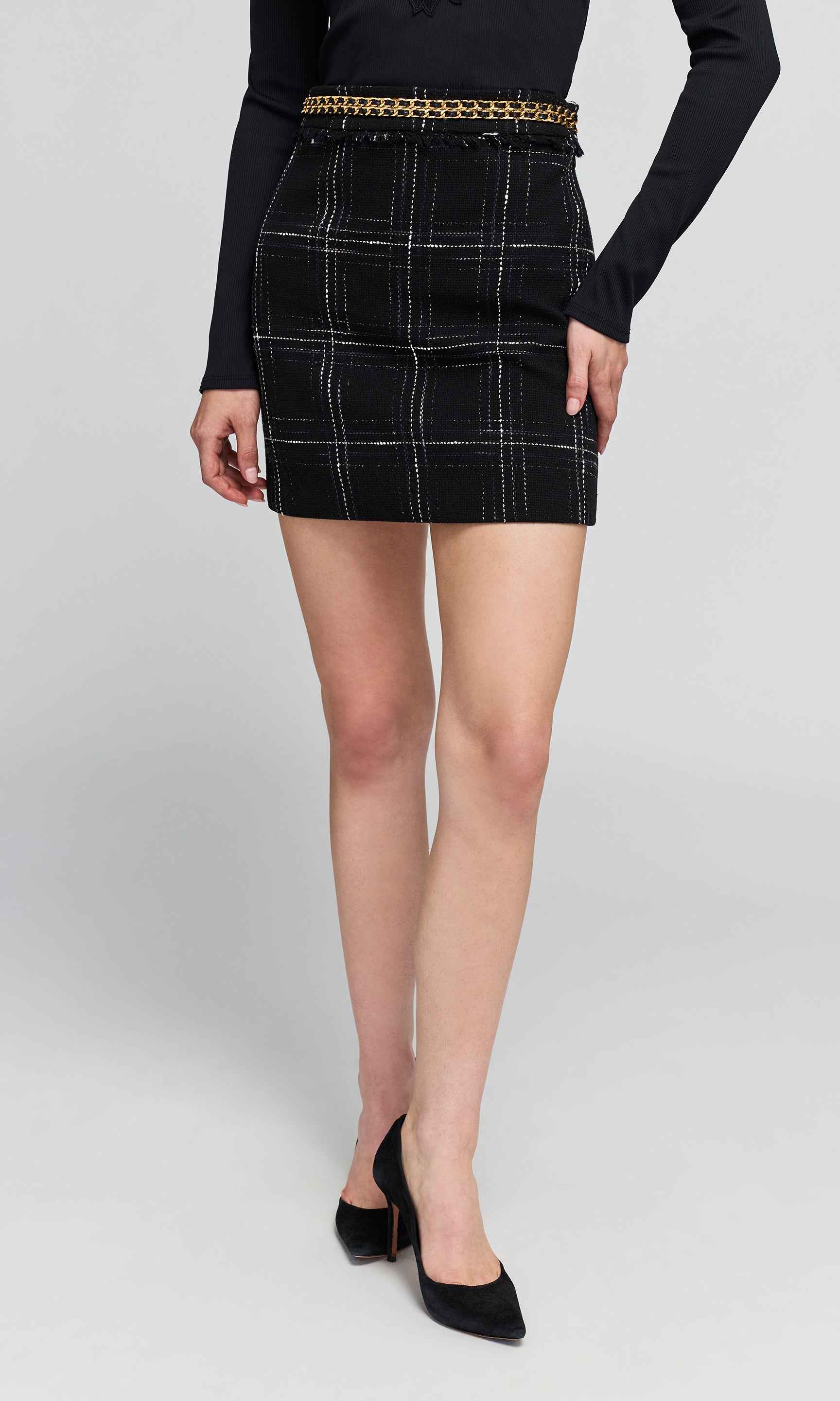 Model wearing a black high-waisted plaid mini skirt with white windowpane lines, adorned with a gold chain belt, a black long-sleeve top.