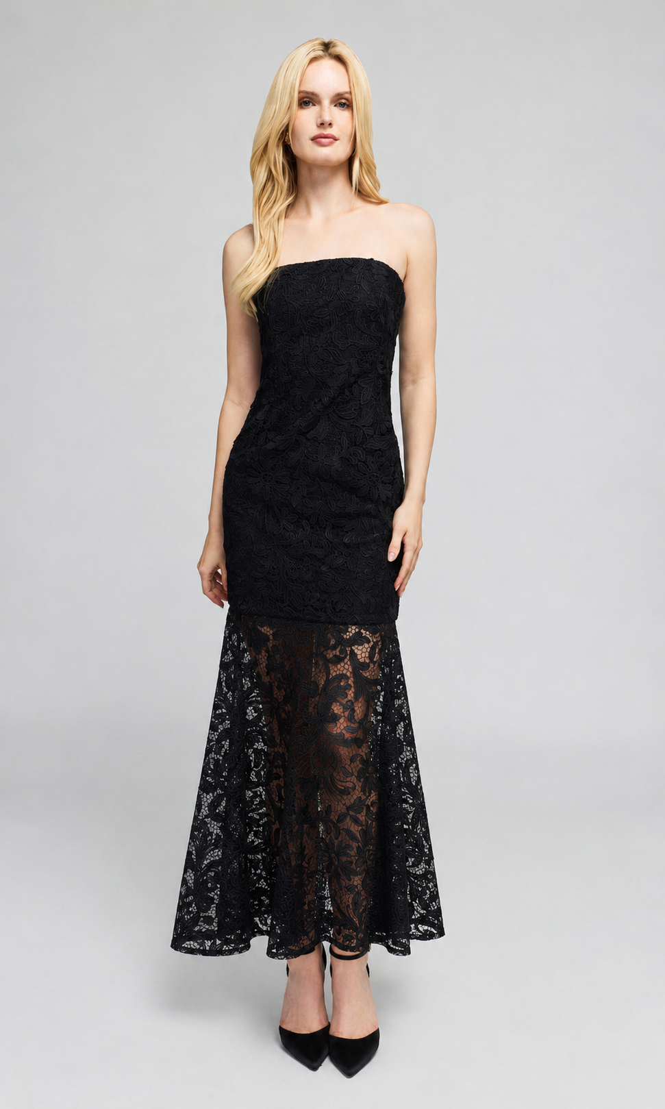 Model wears a strapless black lace maxi dress with a fitted bodice and a flared lace skirt, paired with black pointed-toe heels.