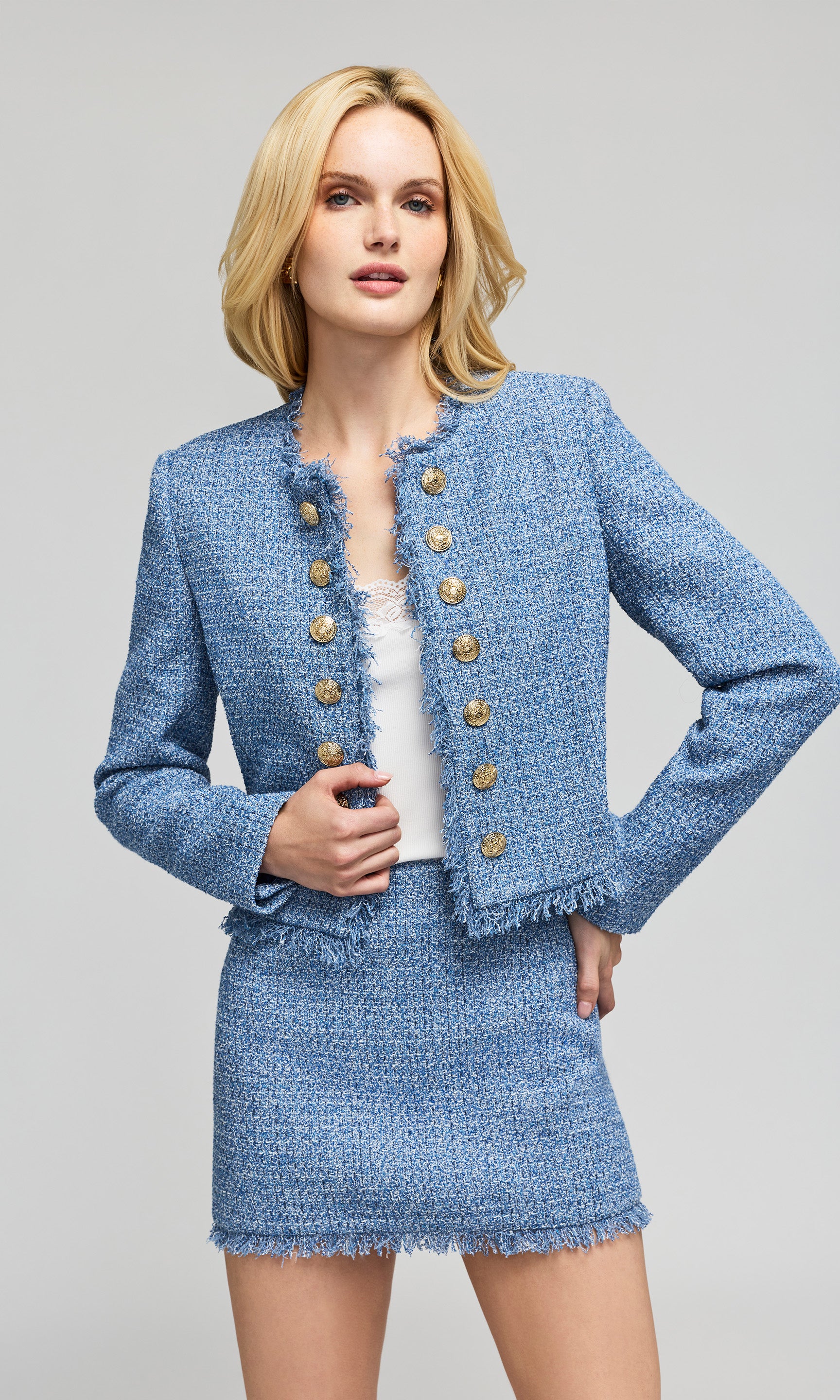 Model wearing a light blue tweed jacket and matching mini skirt with gold buttons, on a plain background.