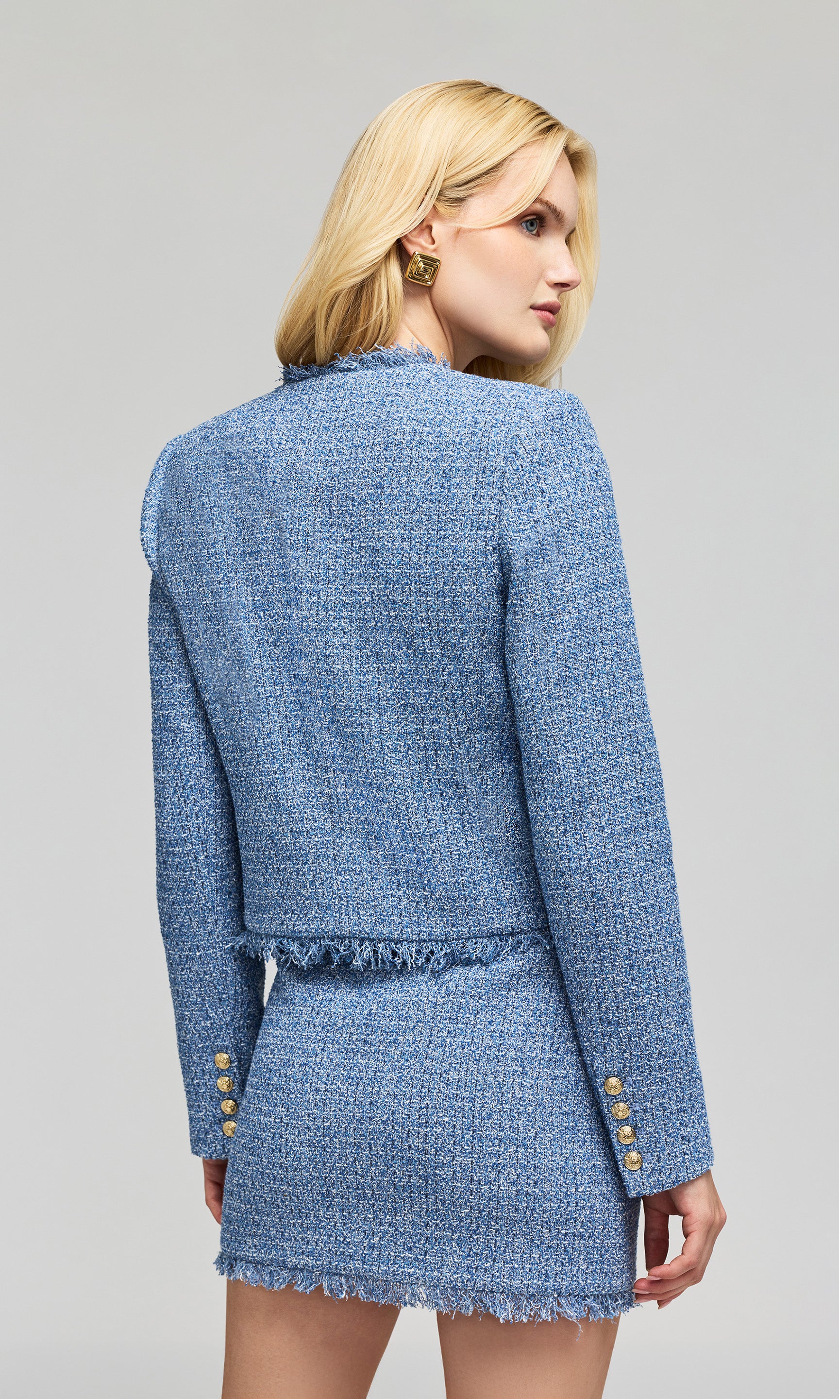 Back view of model wearing a light blue tweed jacket and matching mini skirt with gold buttons, on a plain background.