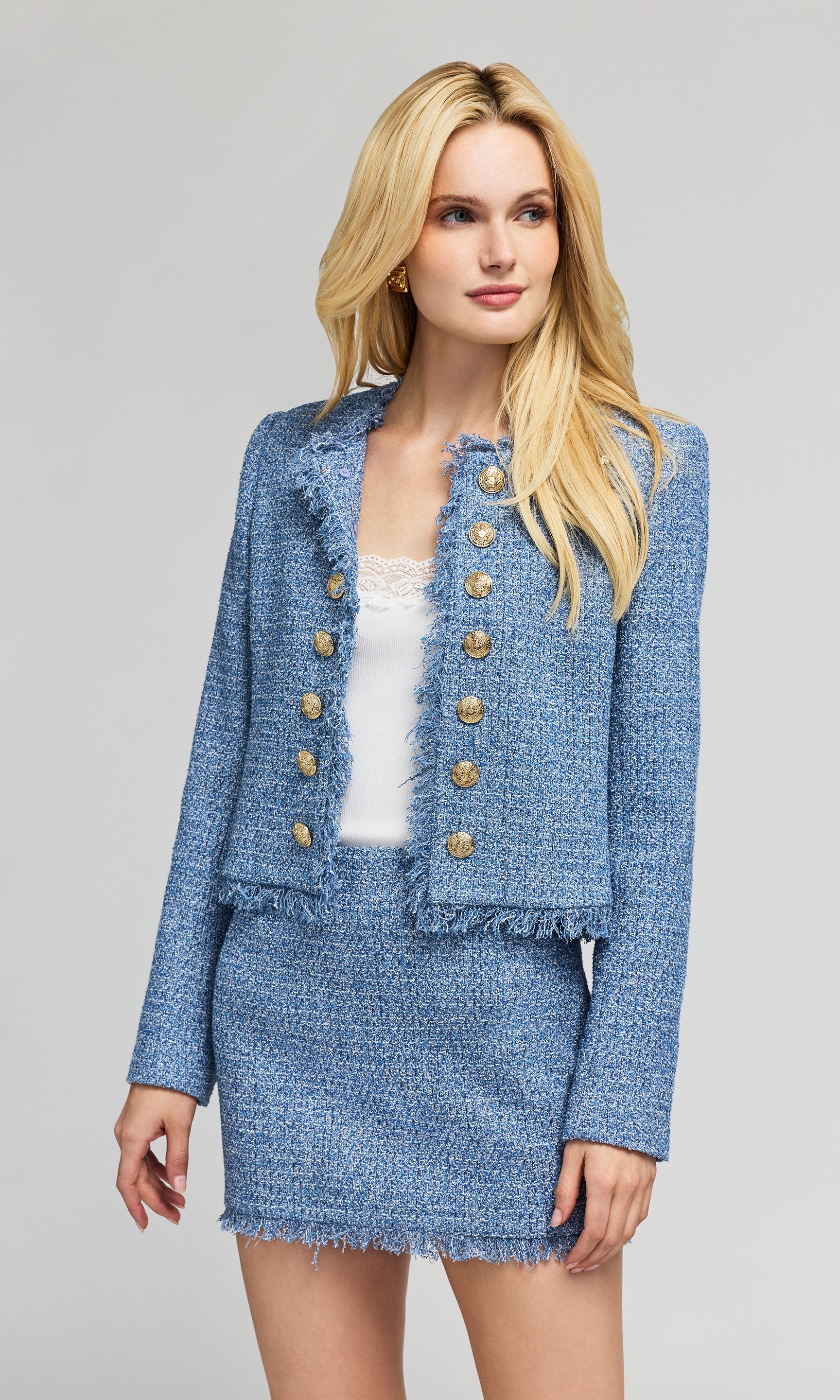 3/4 view of model wearing a light blue tweed jacket and matching mini skirt with gold buttons, on a plain background.