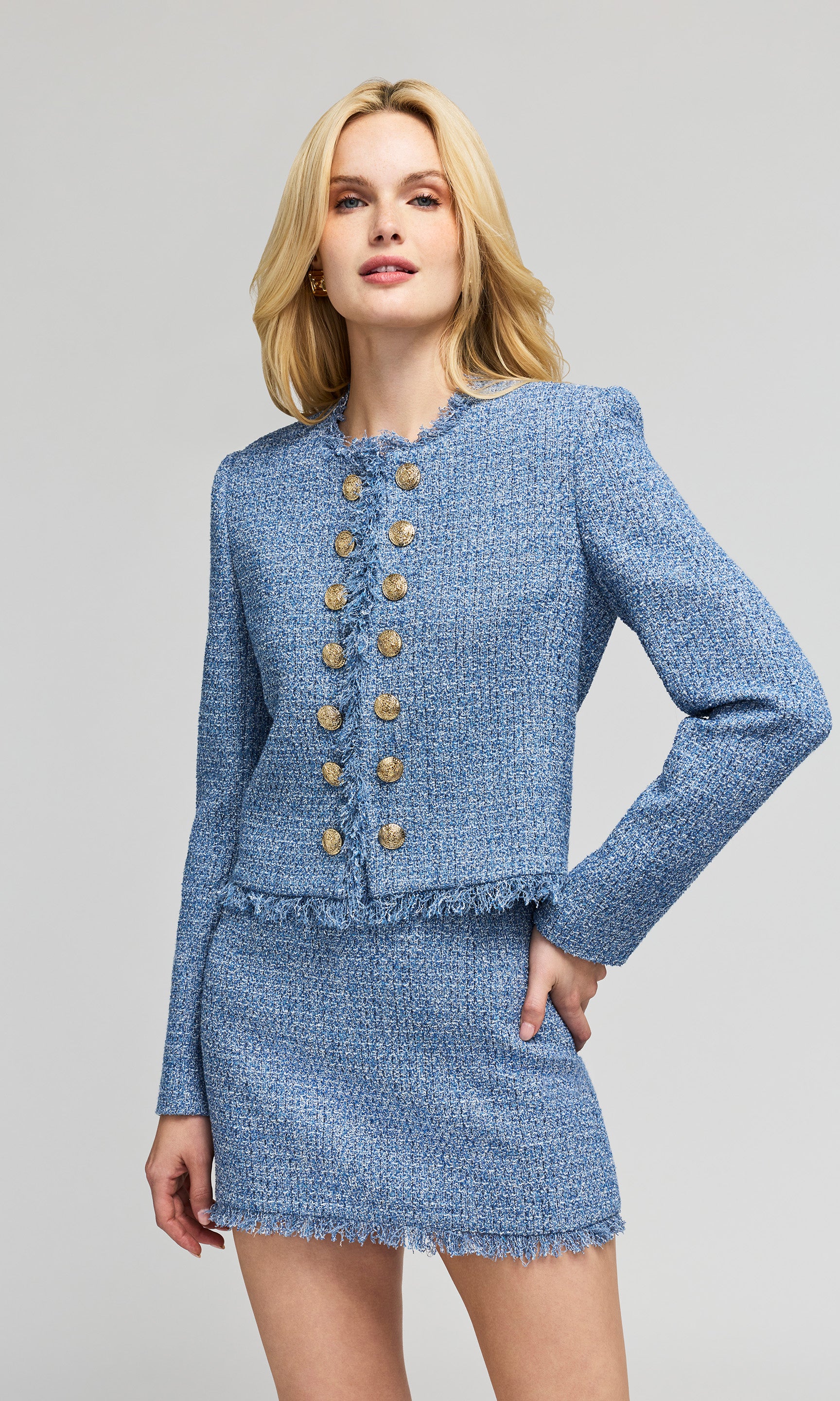 Model wearing a light blue tweed jacket and matching mini skirt with gold buttons, on a plain background.