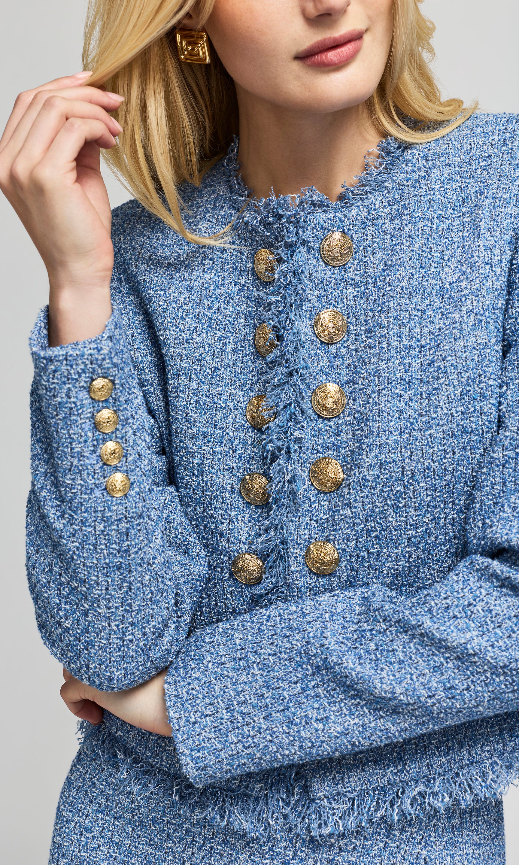 A close-up of a model wearing a blue tweed jacket with prominent gold buttons on the front and cuffs.