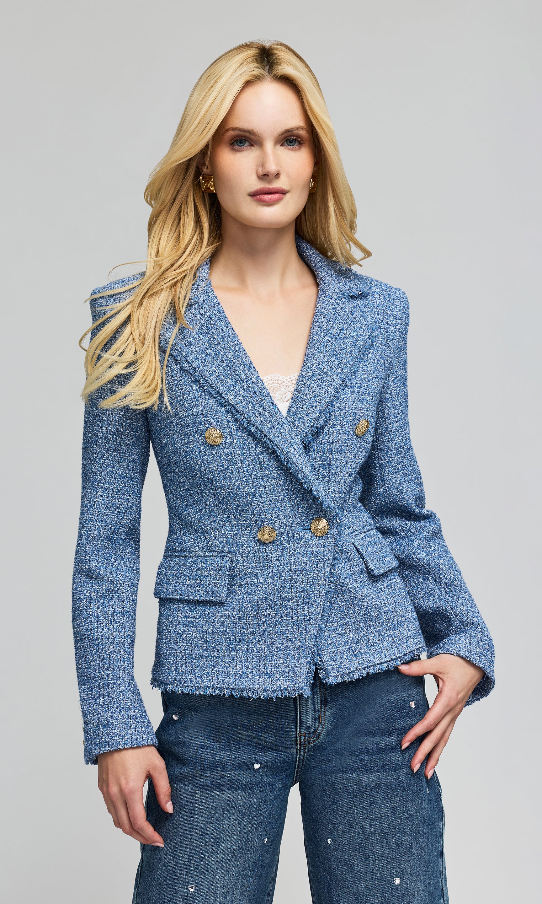 Model wears a tweed blue double-breasted blazer with gold buttons, paired with blue jeans, against plain background.