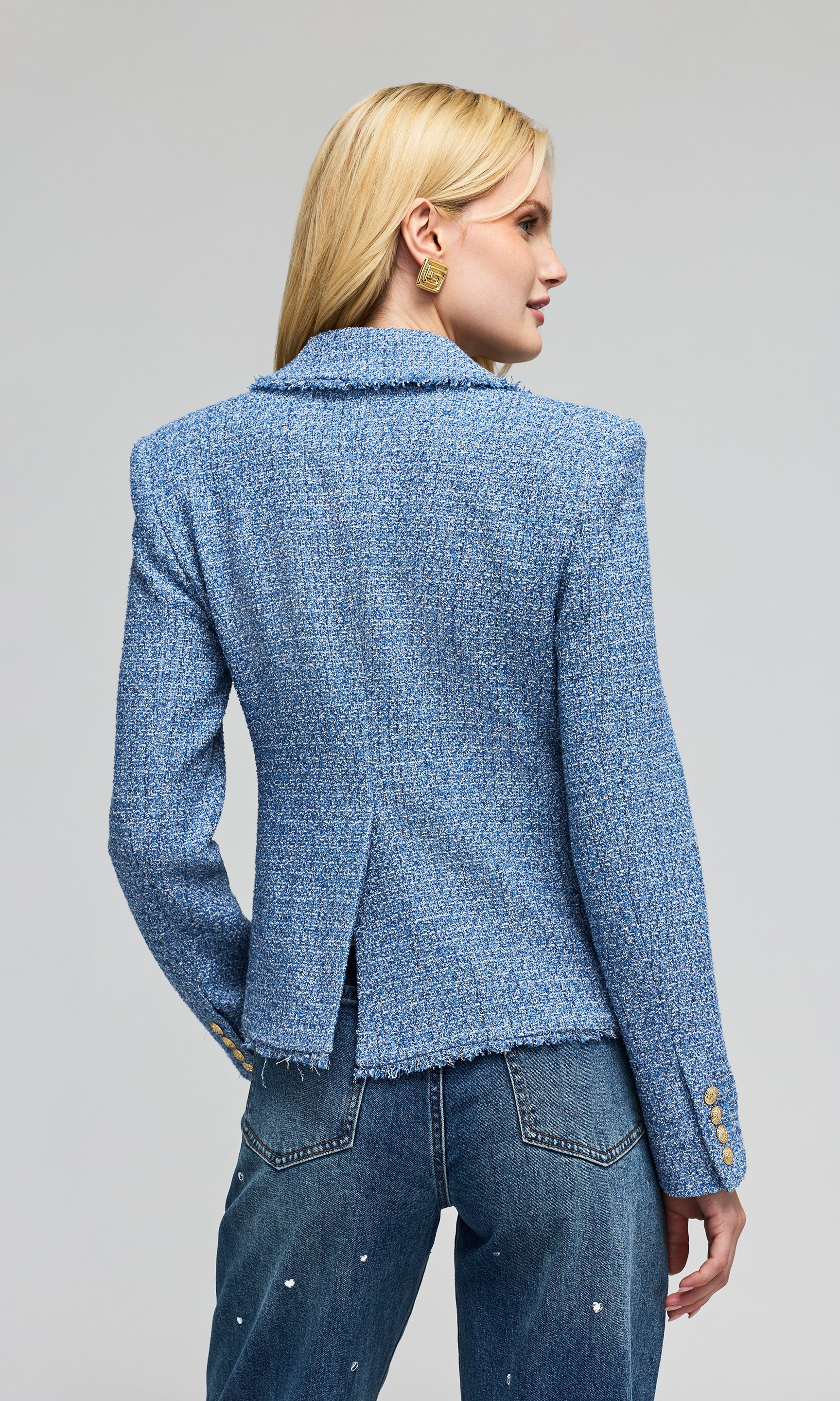 Back view of model wears a tweed blue double-breasted blazer with gold buttons, paired with blue jeans, against plain background.
