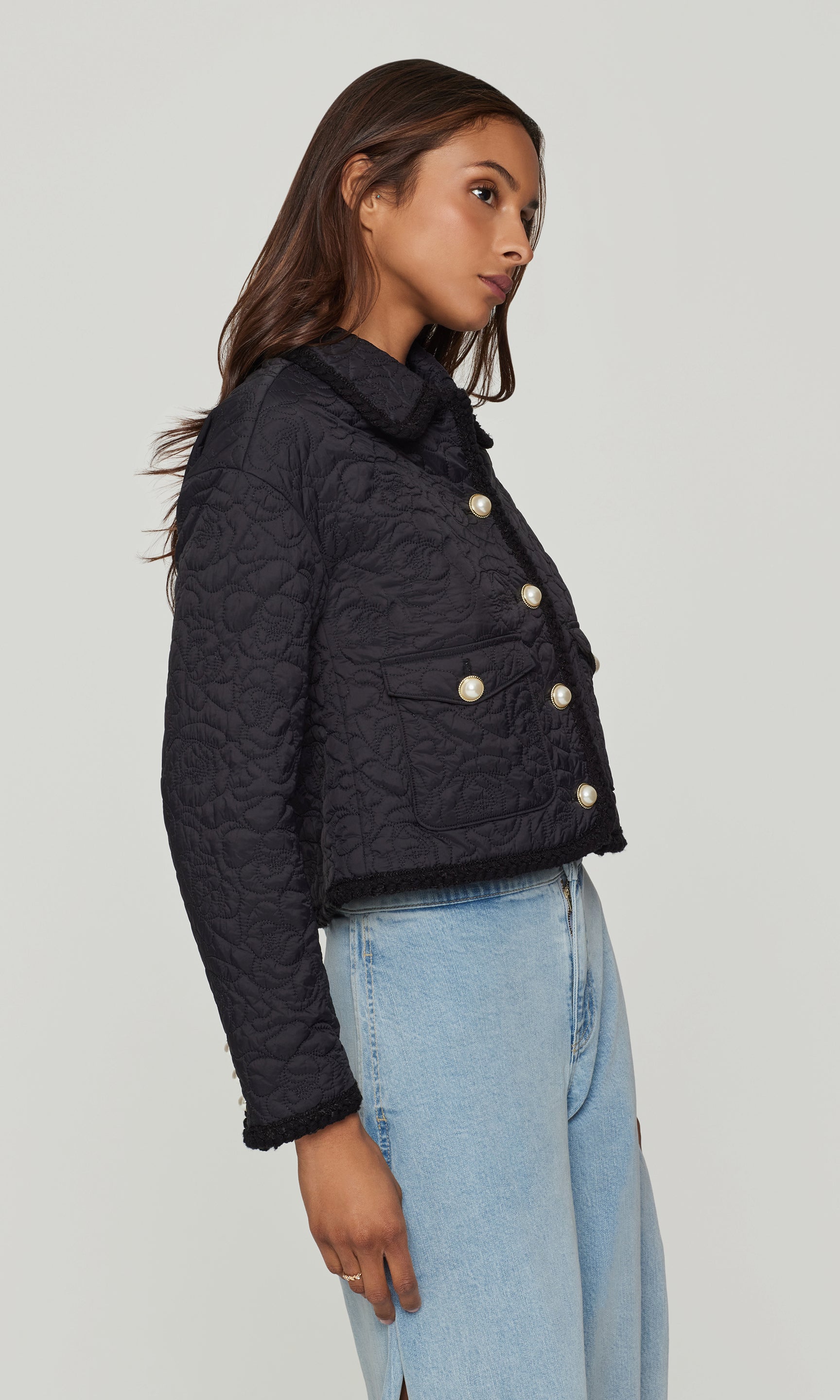 Side view of woman wearing a black quilted jacket with white pearl buttons on a plain background