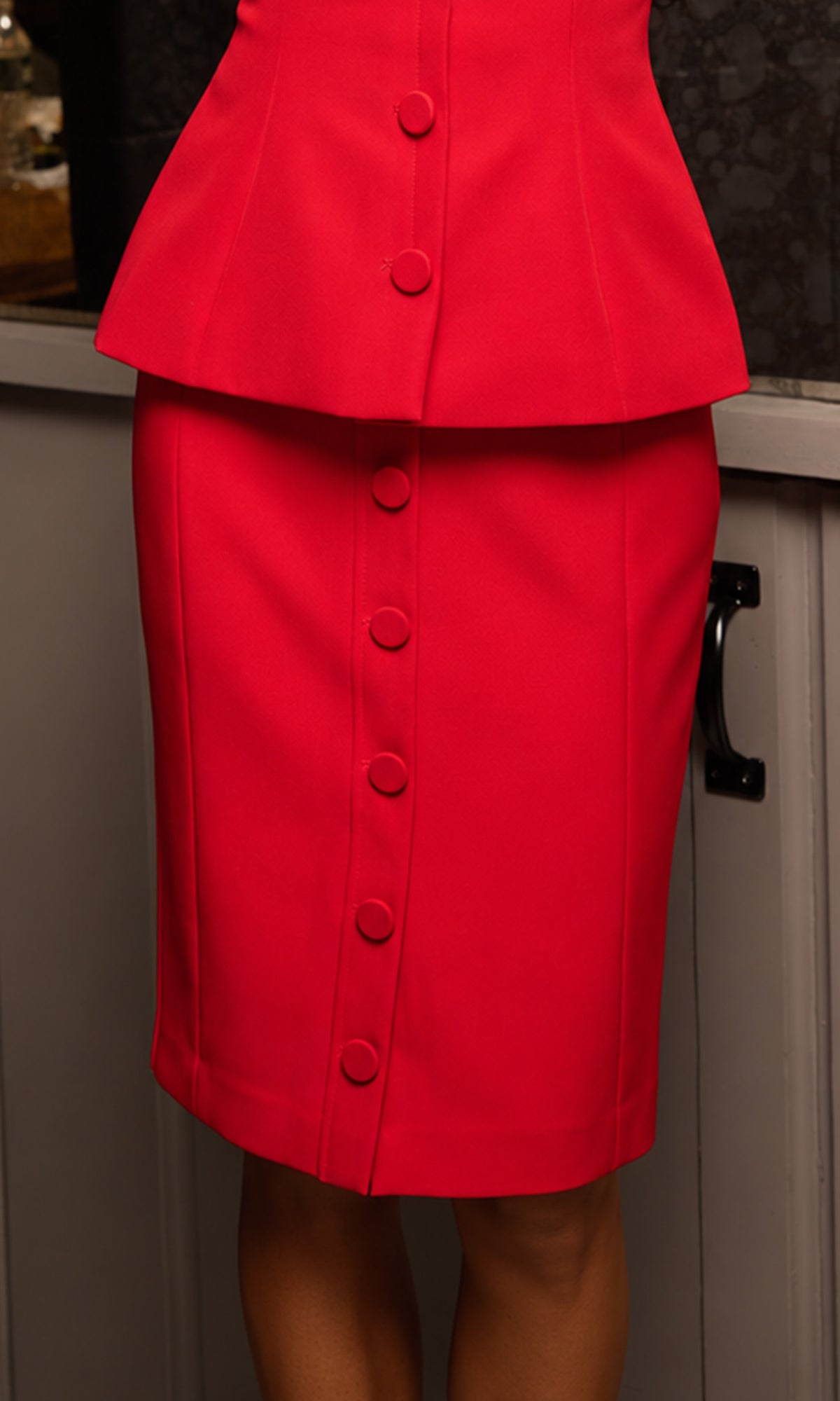 A bright red two-piece skirt suit featuring a structured peplum top with a vertical row of buttons and a matching pencil skirt with a central button placket.