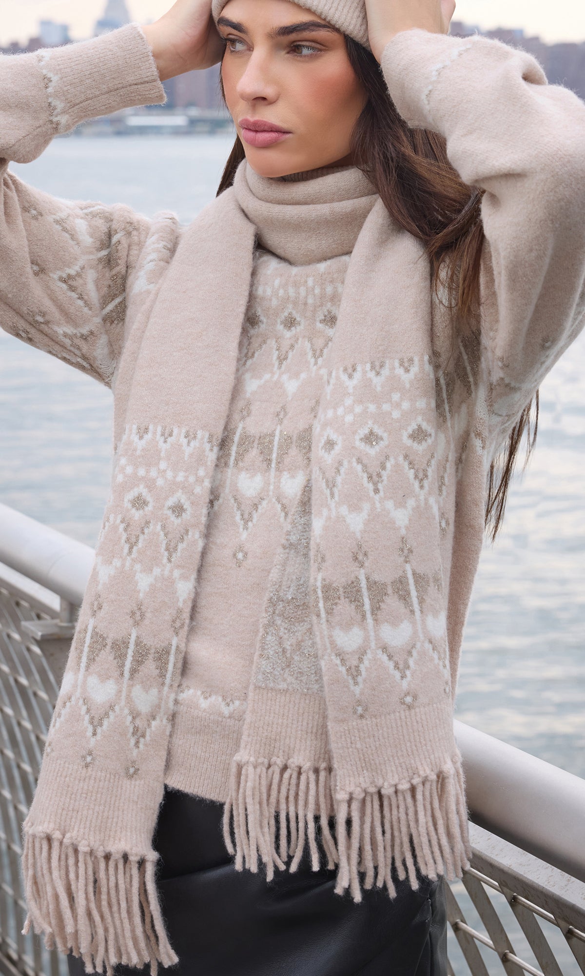 A woman wearing a beige knit sweater and matching scarf with a white Fair Isle pattern, standing outdoors near a railing with water in the background.