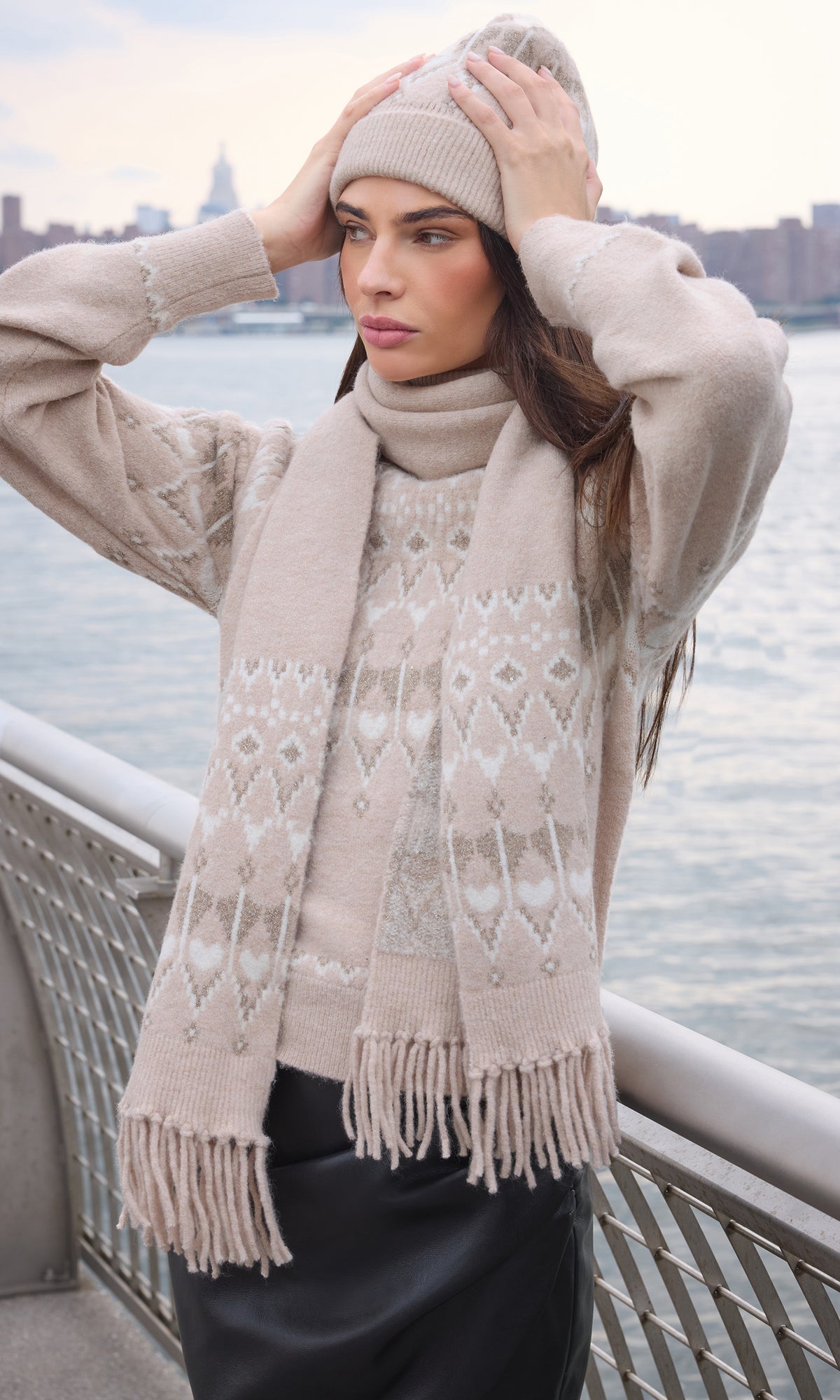 A woman wearing a beige knit sweater, matching scarf, and beanie stands on an outdoor balcony with water and a city skyline in the background.