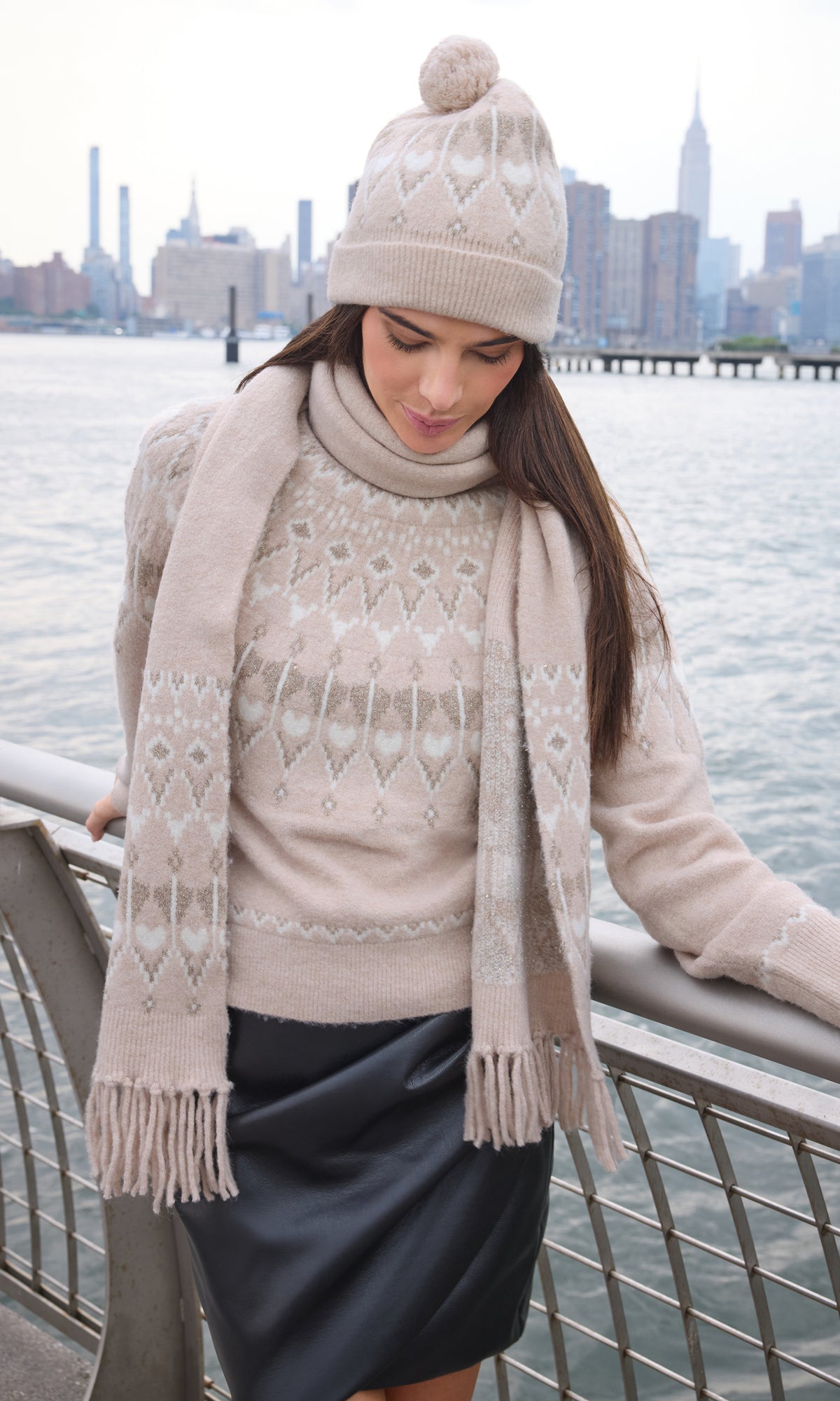 A woman wearing a beige and white patterned knit sweater, matching beanie with pom-pom, and a long fringed scarf, standing by a waterfront railing with a city skyline in the background.
