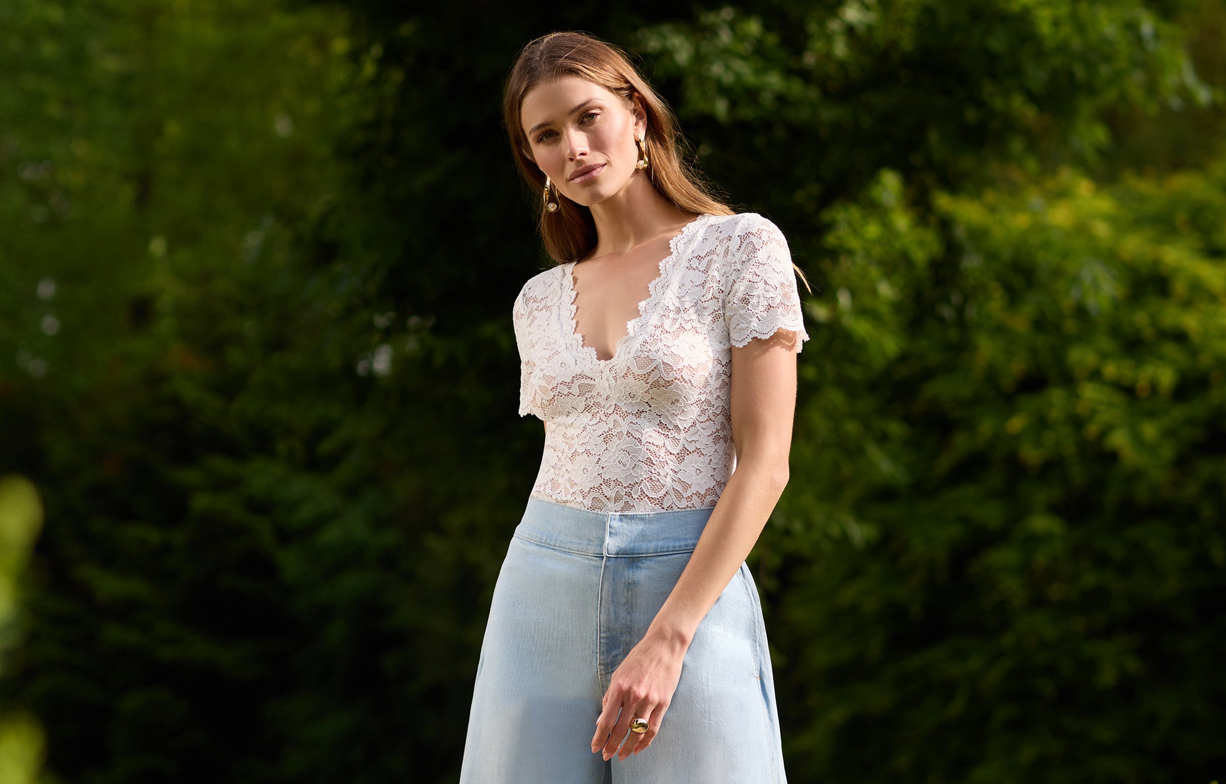 side view of model outdoors in a garden posing wearing the Abigail lace top in white with blue jeans