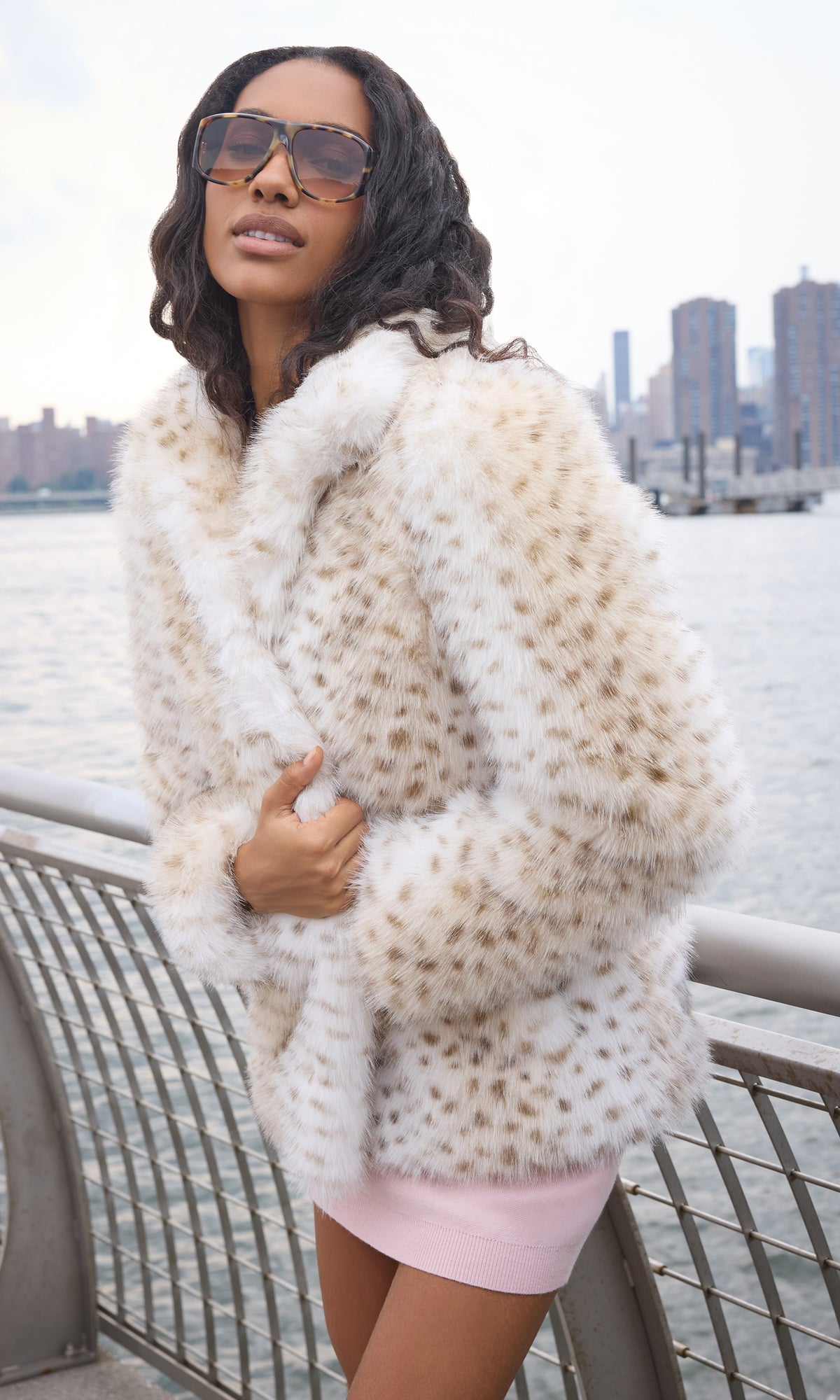 A woman wearing a cream and brown faux fur jacket with a leopard-like pattern, standing outdoors near a waterfront railing with a city skyline in the background.