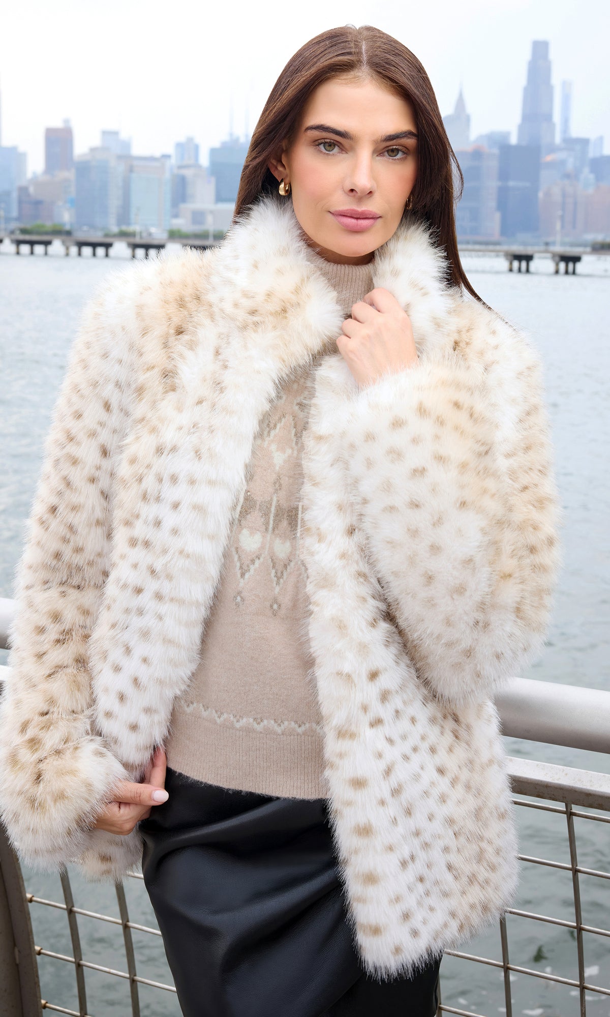 A woman wearing a cream and beige spotted faux fur coat over a beige sweater and black skirt, standing outdoors with a city skyline and water in the background.