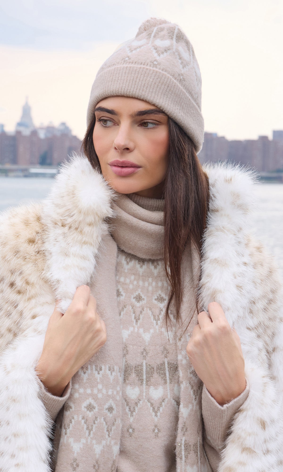 A woman wearing a beige knit beanie, matching knit sweater, and a cream faux fur coat with a patterned border, standing outdoors with a blurred city skyline and water in the background.